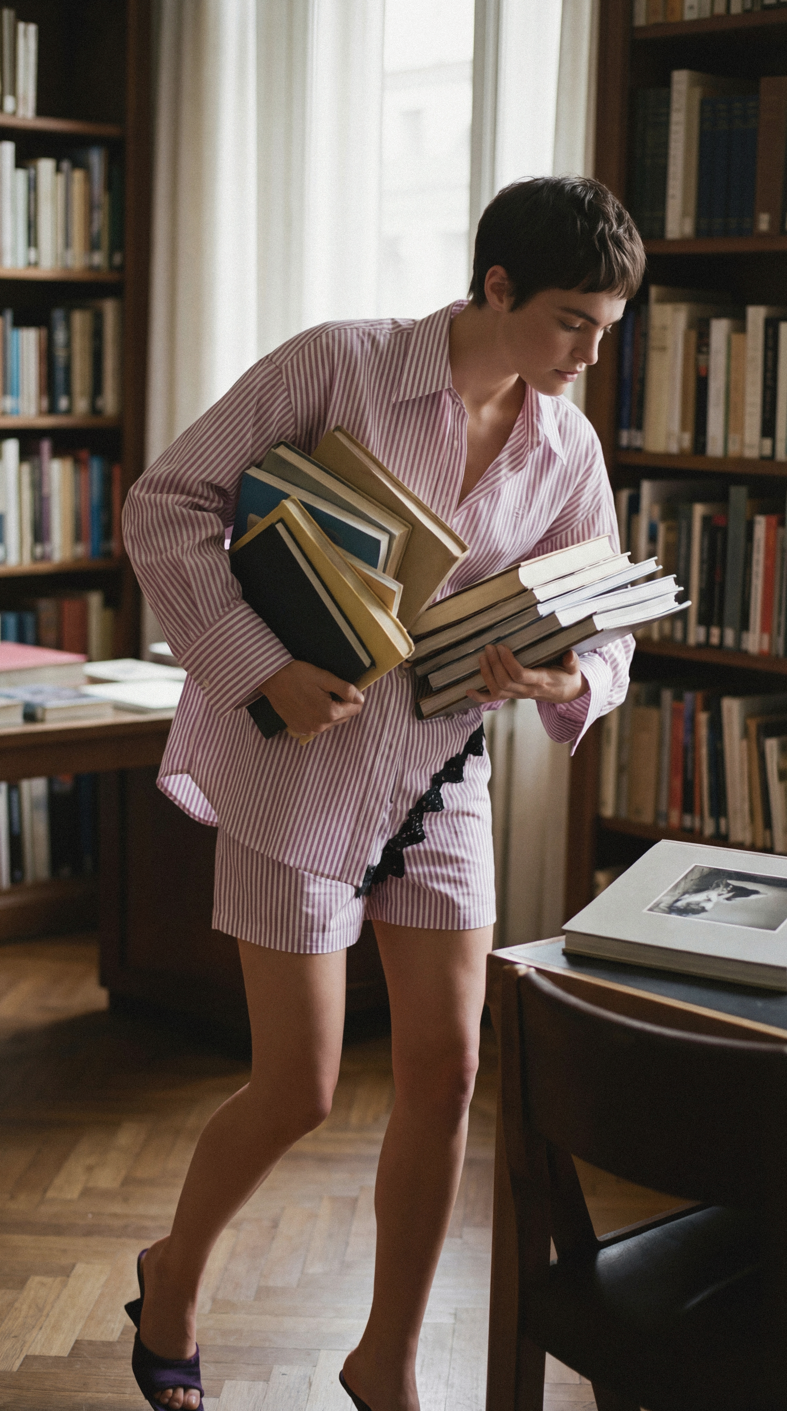 Person in pajamas carrying multiple books in a room filled with bookshelves and a table with a large photo book.