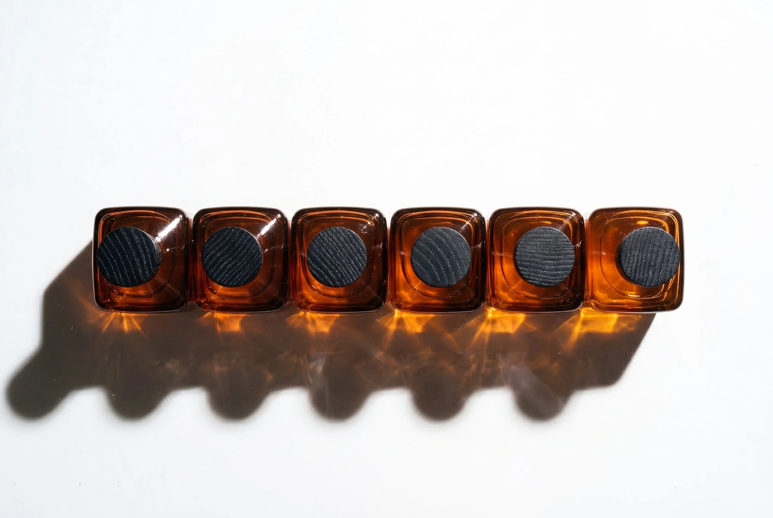 Six amber-colored glass bottles with black, textured caps are lined up in a row on a white surface, casting shadows on the surface.