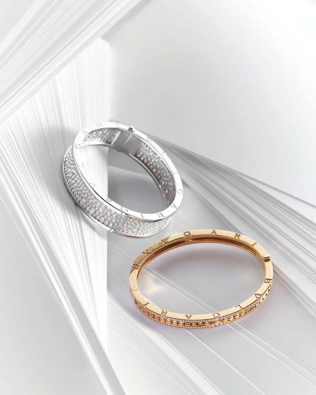 Two luxury Bvlgari bracelets, one silver with diamonds and one gold, resting on white textured background.