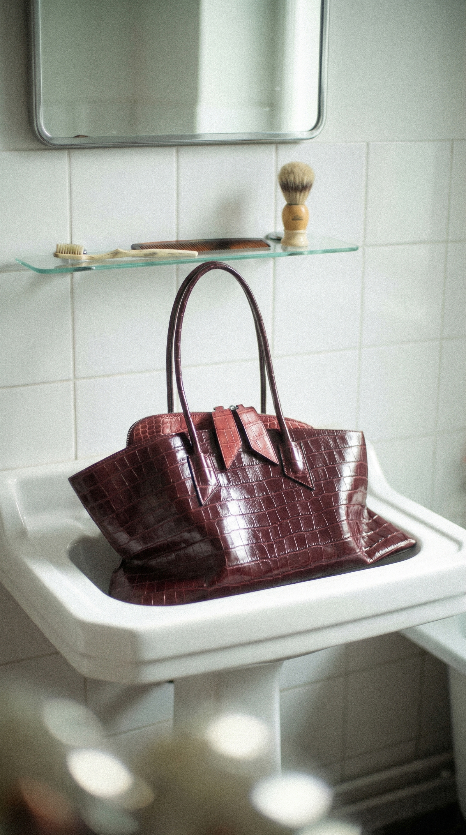 A burgundy crocodile-embossed leather handbag placed on a white bathroom sink, with a shelf above holding a shaving brush, a comb, and a toothbrush.