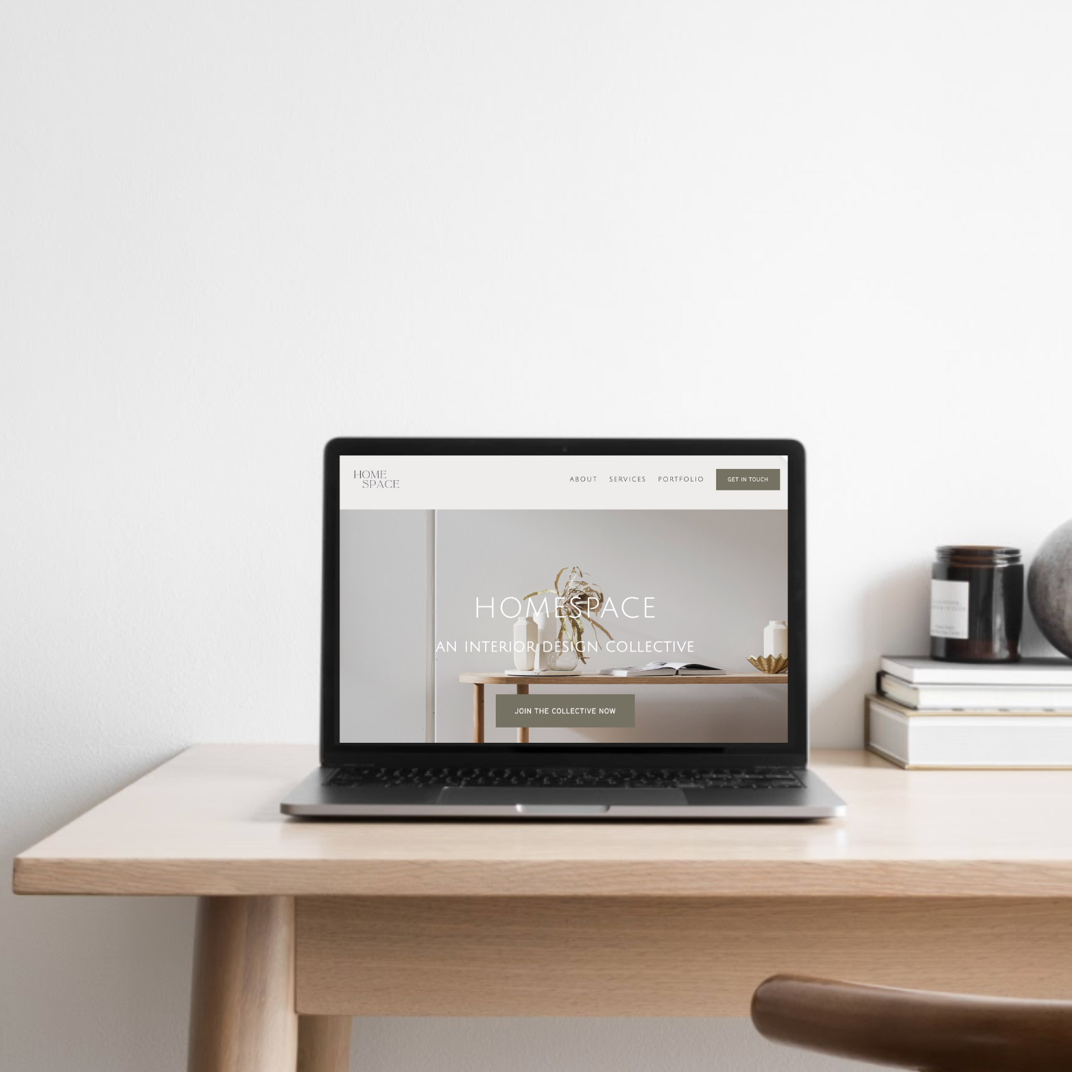 A laptop on a wooden desk displaying a home interior design website, with a white wall and decorative items like a stack of books and a jar nearby.