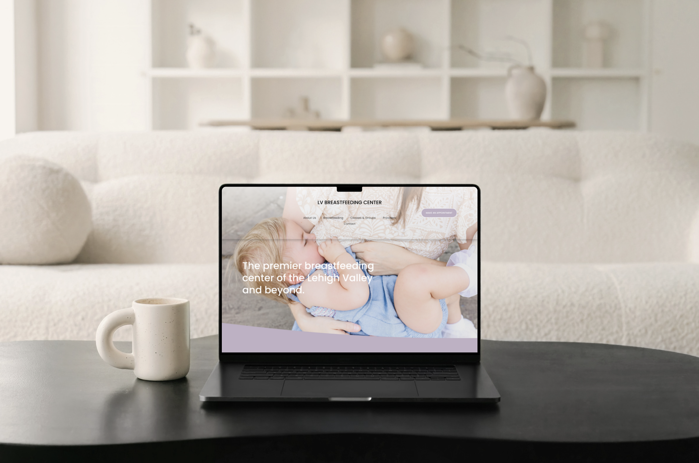 Laptop on a black table displaying a website for an LV Breastfeeding Center with a background photo of a woman breastfeeding a small child. Next to the laptop is a white mug. In the background, there's a white sofa with decorative pillows and white shelving with vases and decor against a light-colored wall.