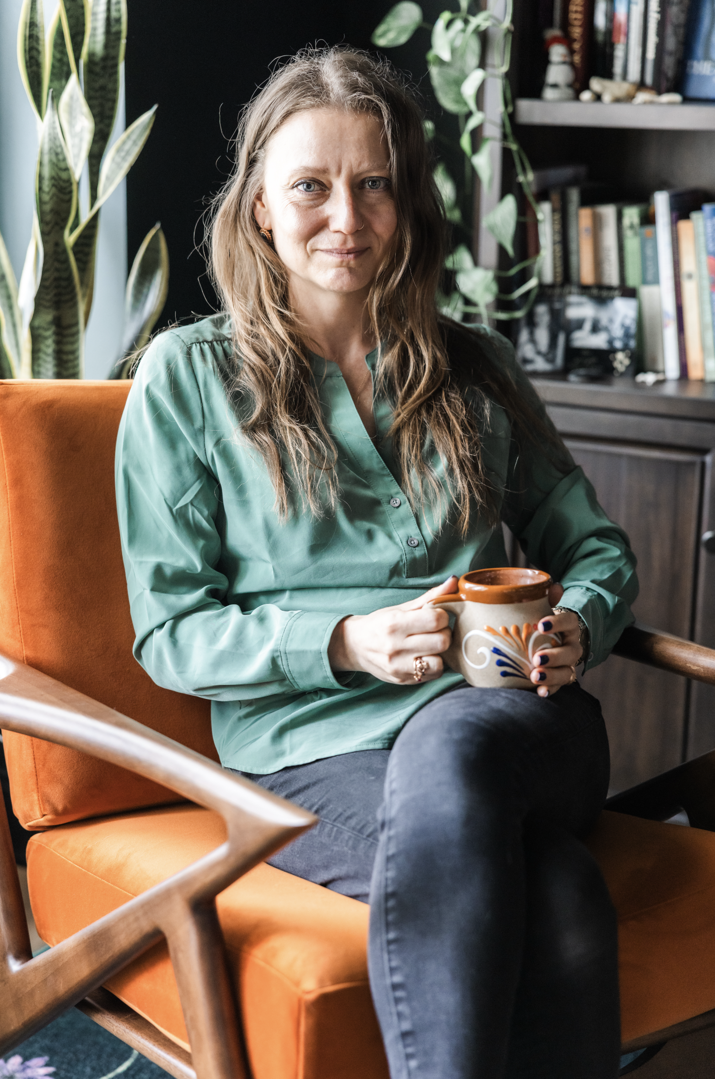 A woman with long wavy hair sitting on an orange chair in a room with green plants and a bookshelf, holding a ceramic mug.