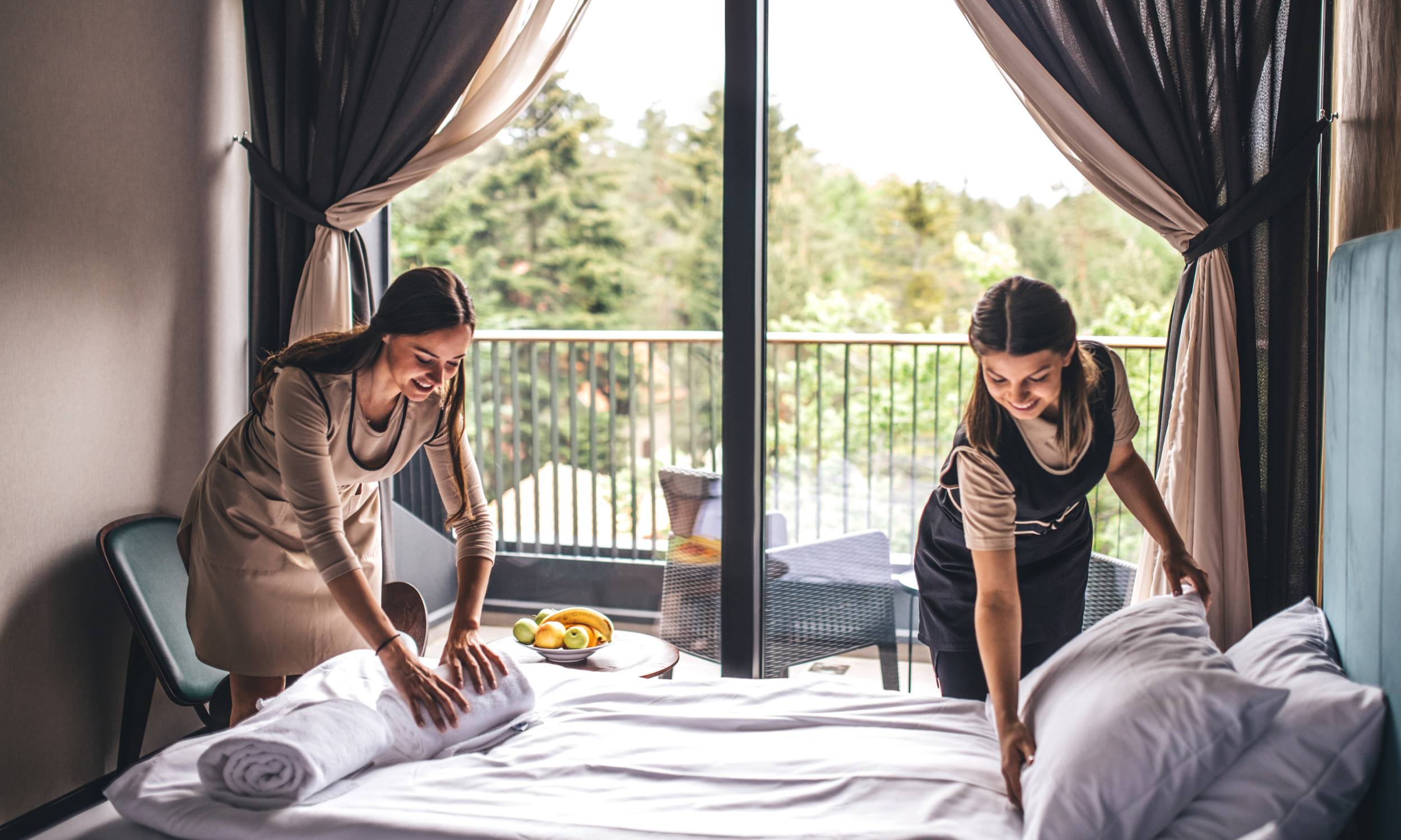 Two hotel housekeepers are making a bed in a room with large sliding glass doors opening to a balcony and a view of green trees. One housekeeper is smiling and lifting a rolled towel, while the other is smoothing a pillow.