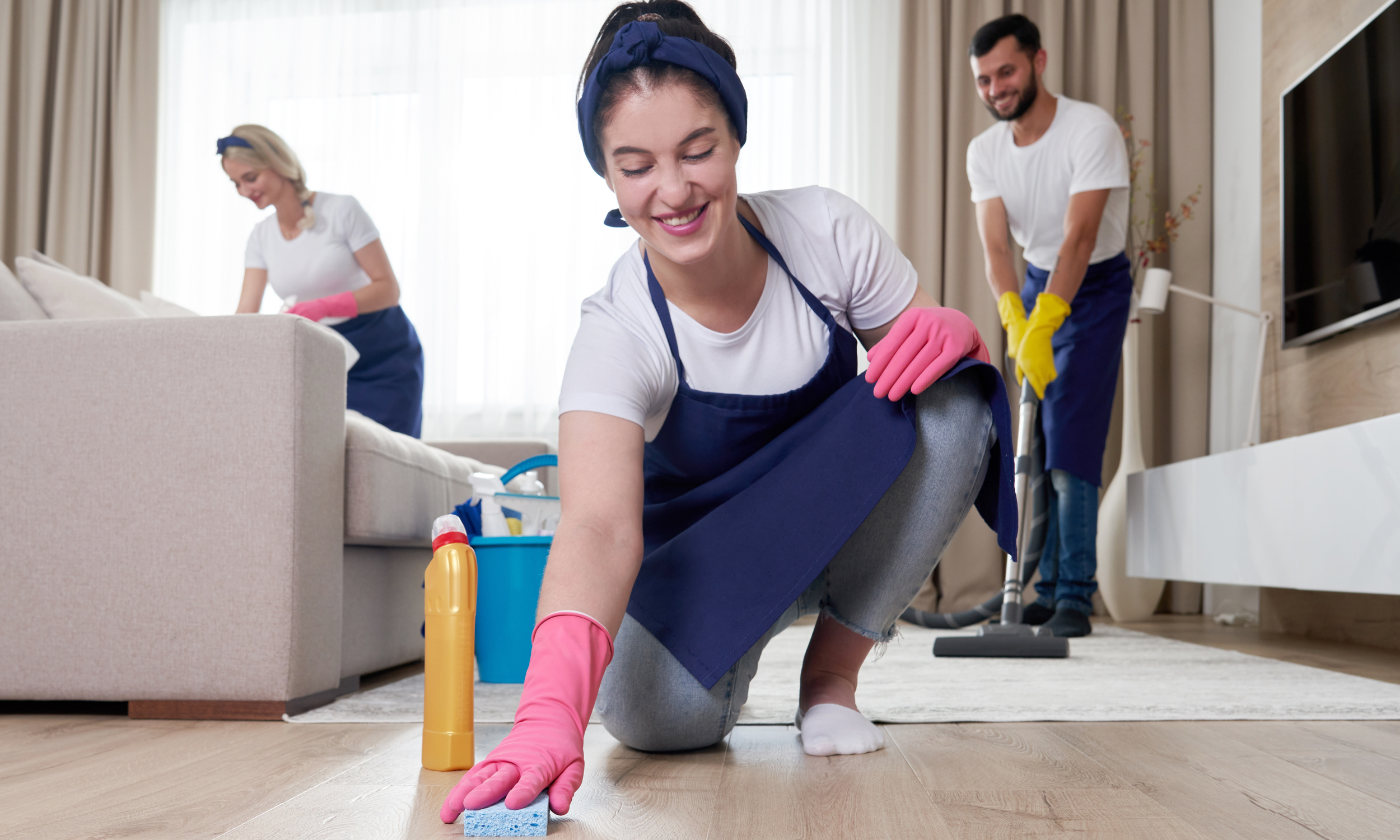 Three people cleaning a living room together, with one woman kneeling on the floor scrubbing, another woman standing near the couch with cleaning supplies, and a man in the background vacuuming.