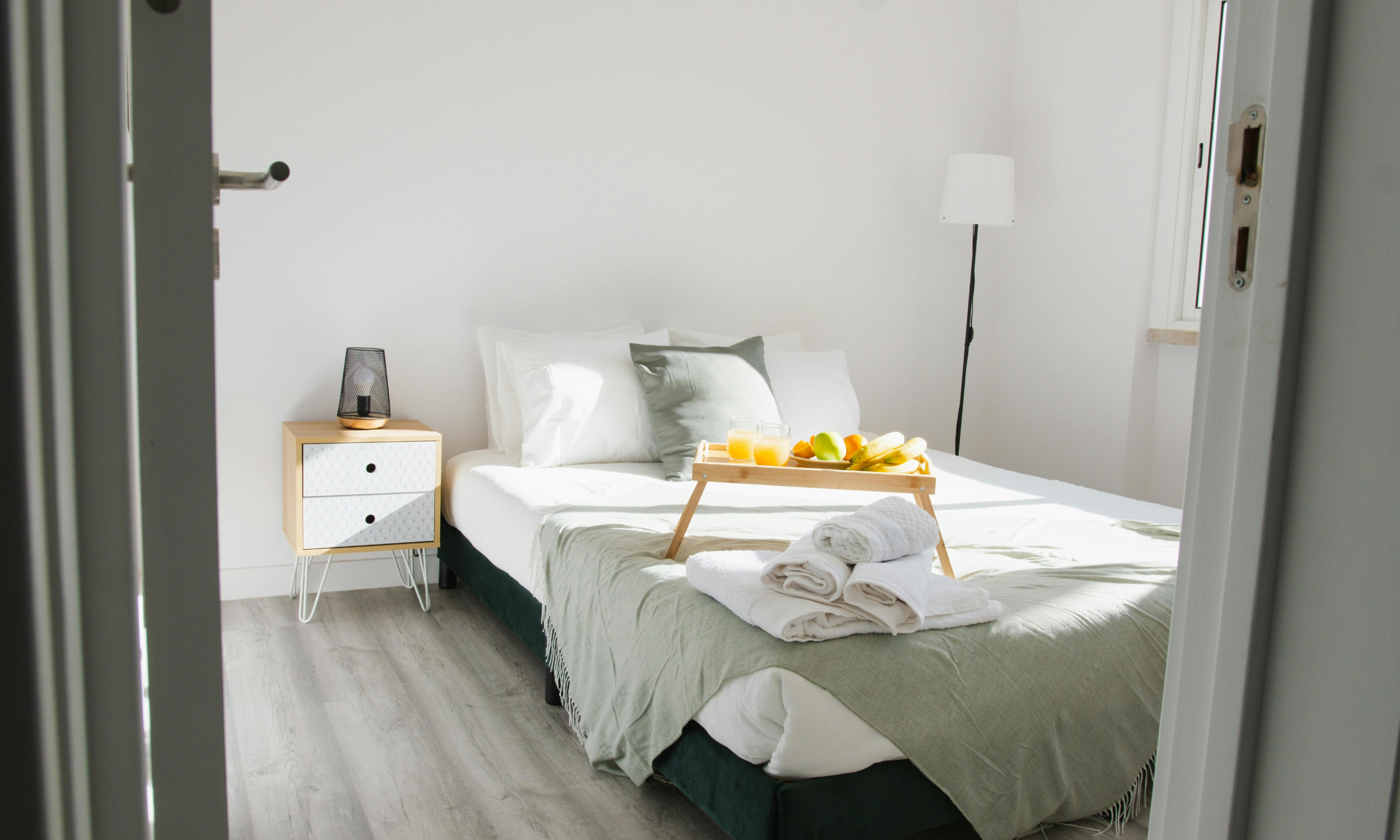 Bright bedroom with a neatly made bed, green and white bedding, and a breakfast tray with orange juice and fruits. There is a nightstand with a lamp, a floor lamp, and a window with natural light.