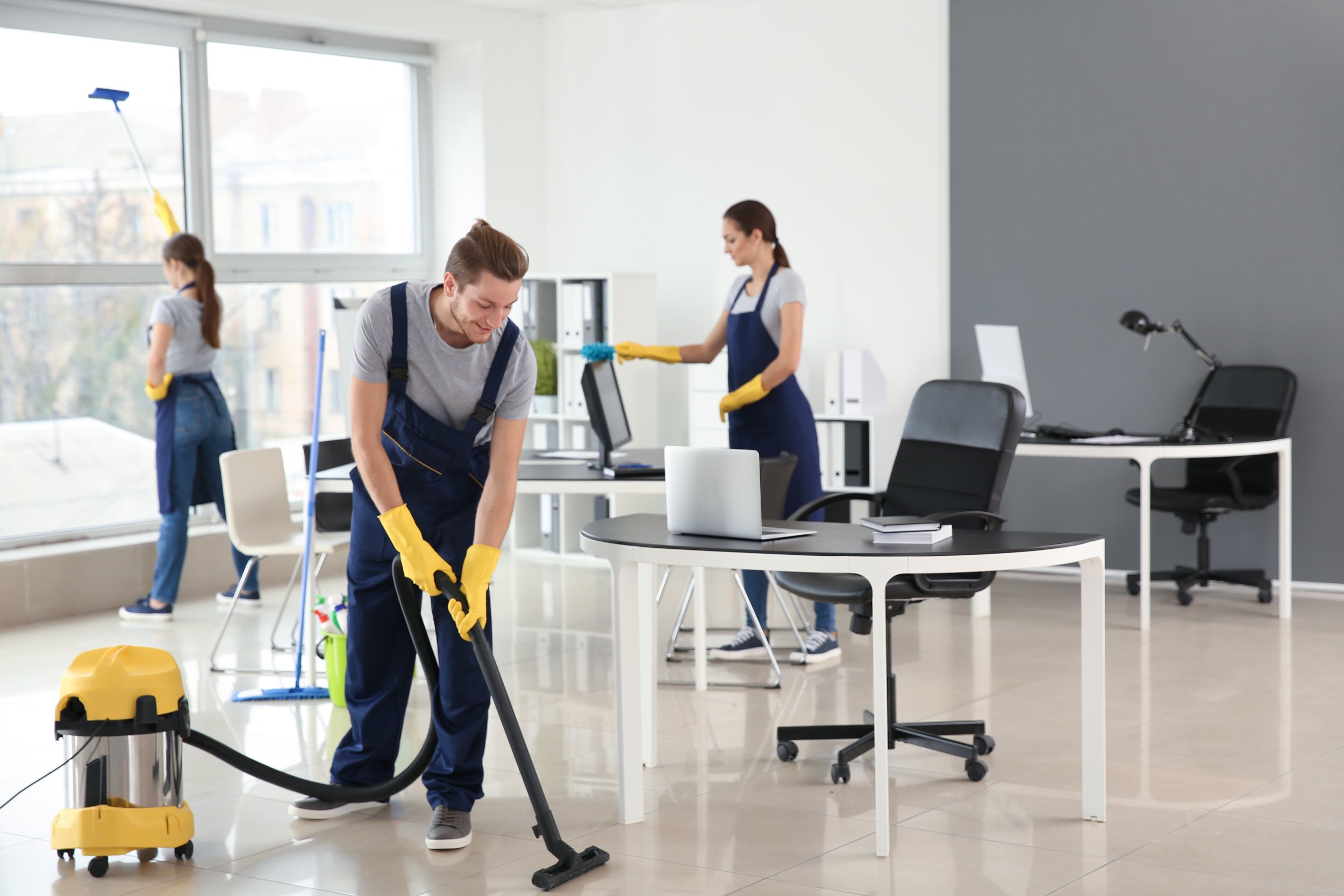 Three people cleaning an office. One person is vacuuming, another is wiping a desk, and a third is mopping near a large window.