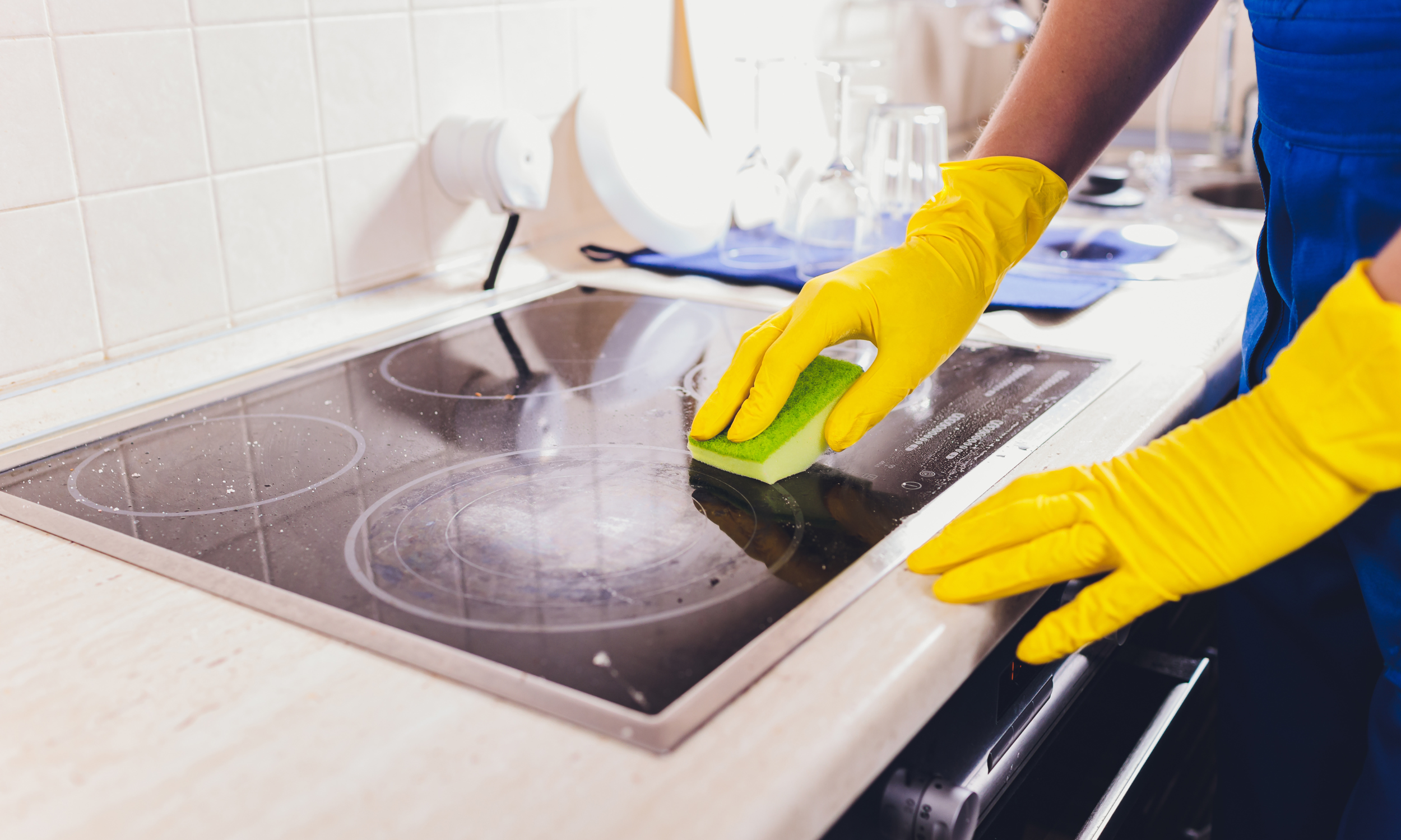 Person wearing yellow gloves cleaning a glass stove top with a green sponge in a kitchen.