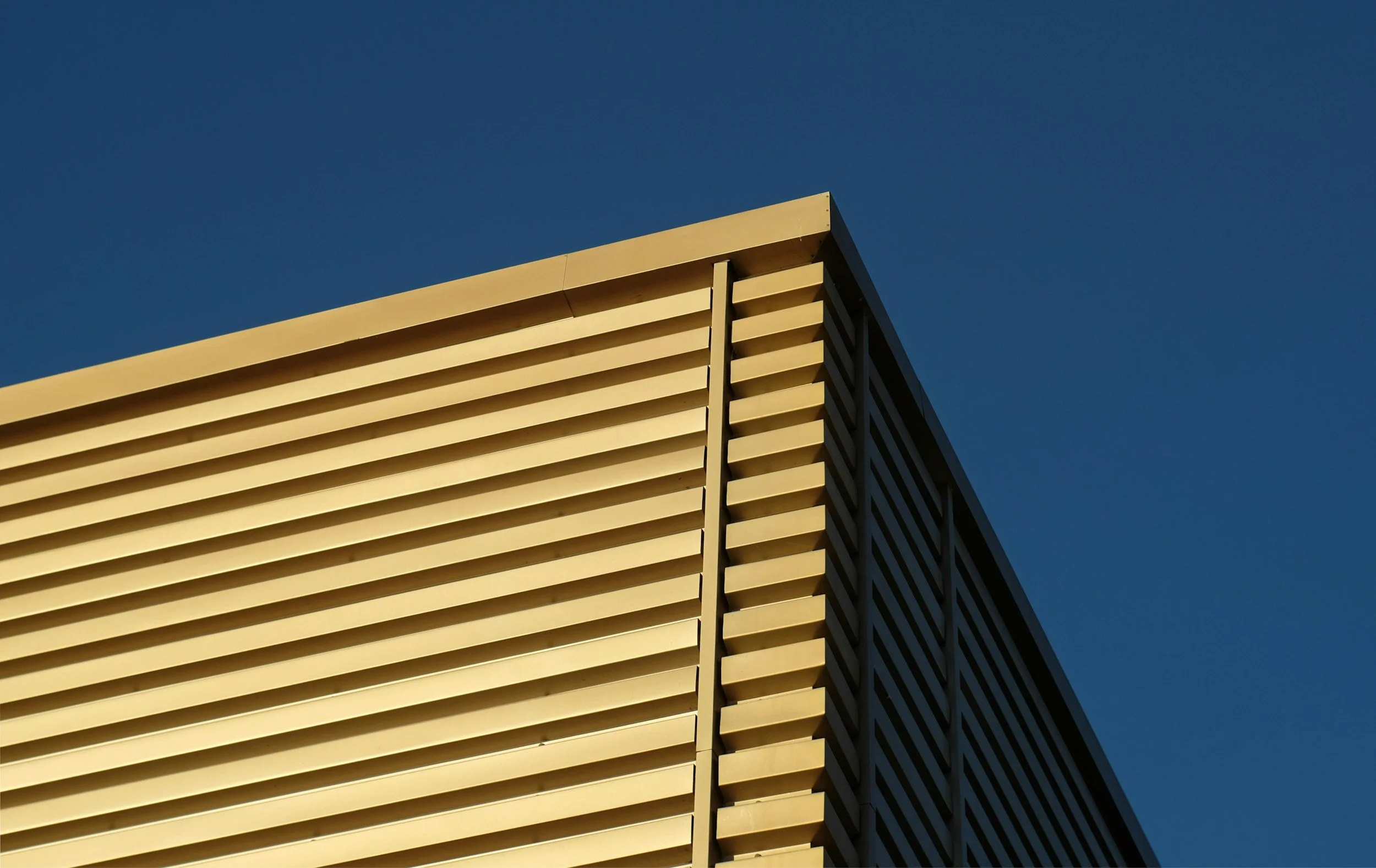 Close-up of a modern building corner with horizontal beige slats against a clear blue sky.