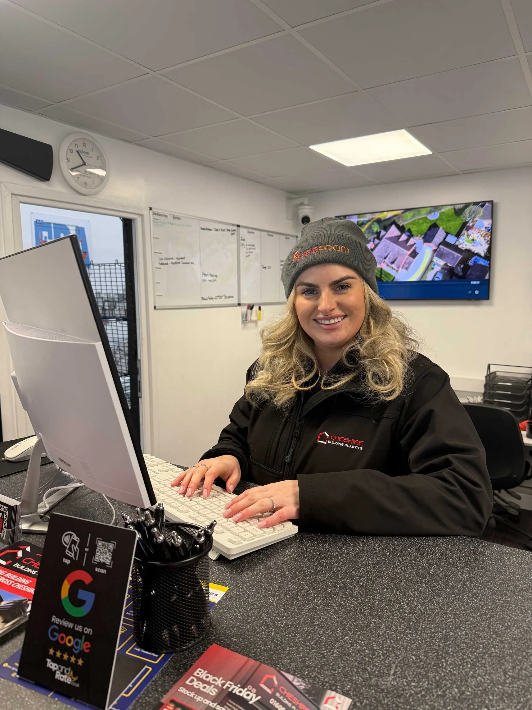 A woman with blonde curly hair, wearing a grey beanie and a black jacket, is sitting at a desk with a computer, smiling at the camera in an office environment. There are promotional materials, pens, and a QR code on the desk.