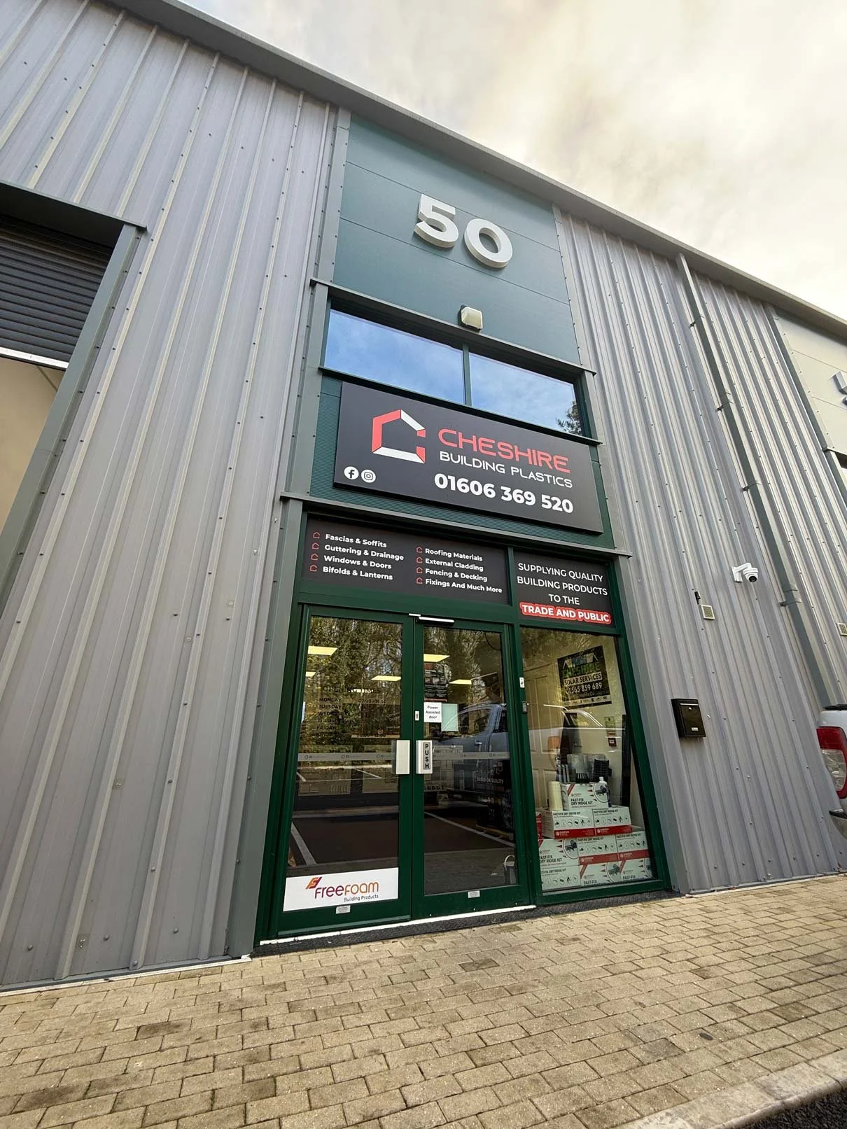 Exterior view of a commercial building with a large sign displaying the name Cheshire Building Plastics and their contact number. The building has a grey metal facade with a glass entrance door. The number 50 is prominently displayed above the sign.