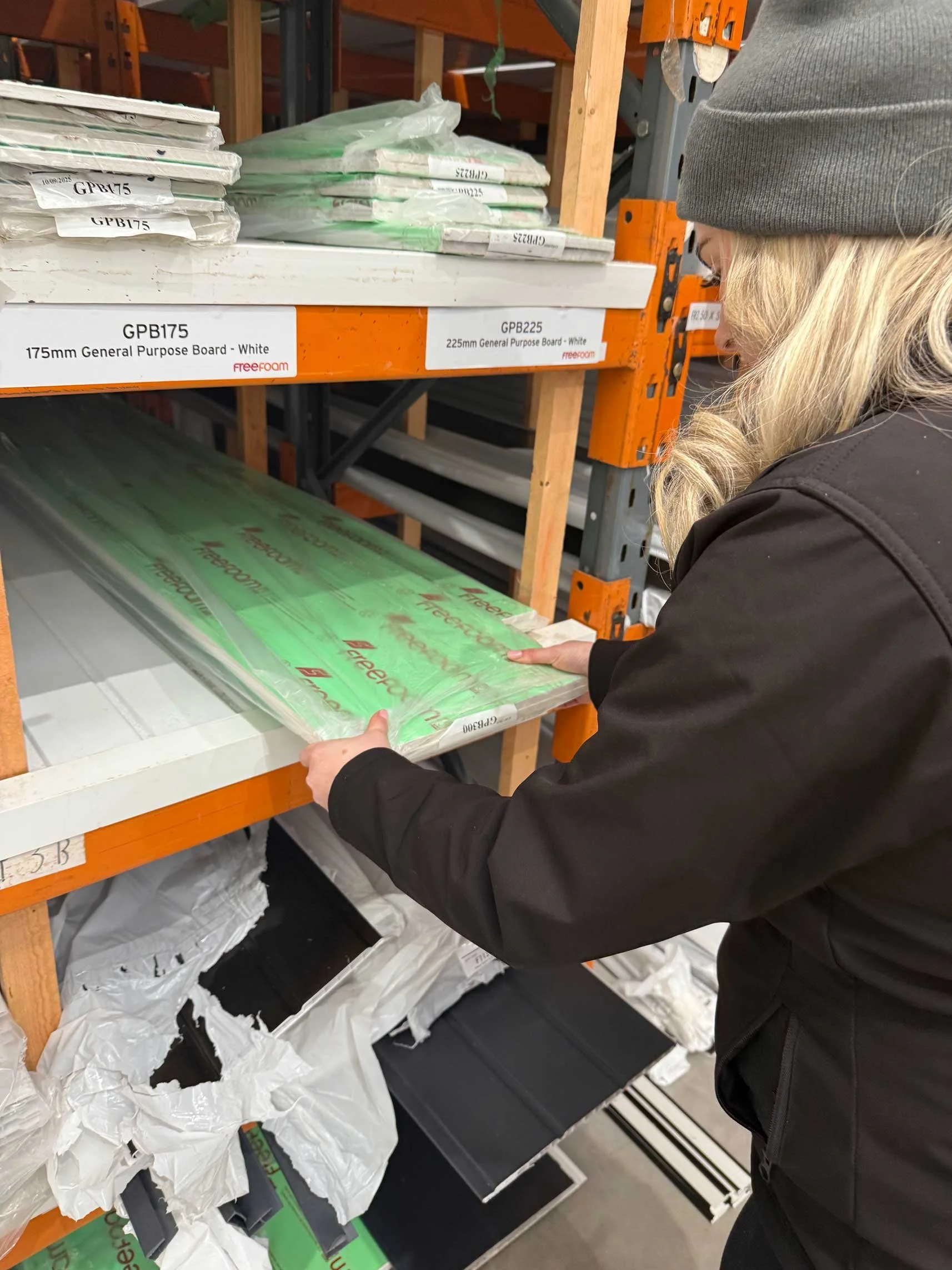 A woman wearing a gray beanie and black jacket at a building supplies store shelf, holding a large sheet of white general purpose board.