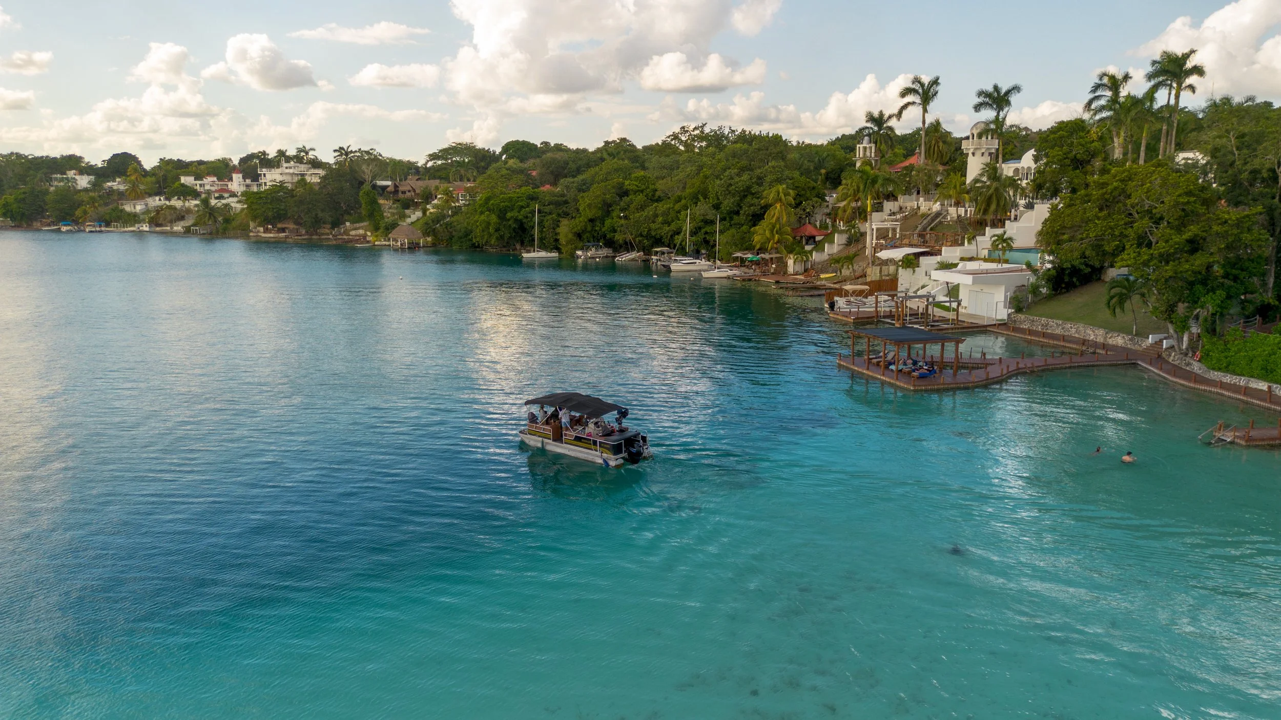 A boat with a canopy floating on clear blue water near a waterfront with white buildings, palm trees, and lush greenery.