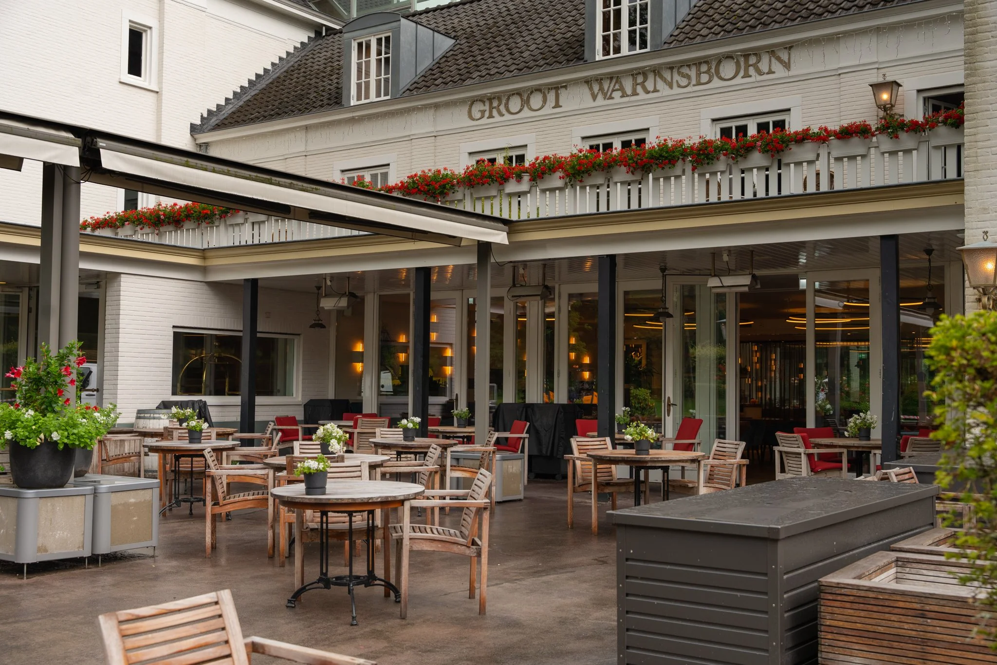 terras met houten tafels en stoelen, bloemenpotten, een clubhuis met balkon en een donkere lucht. Landgoed Groot Warnsborn in Arnhem