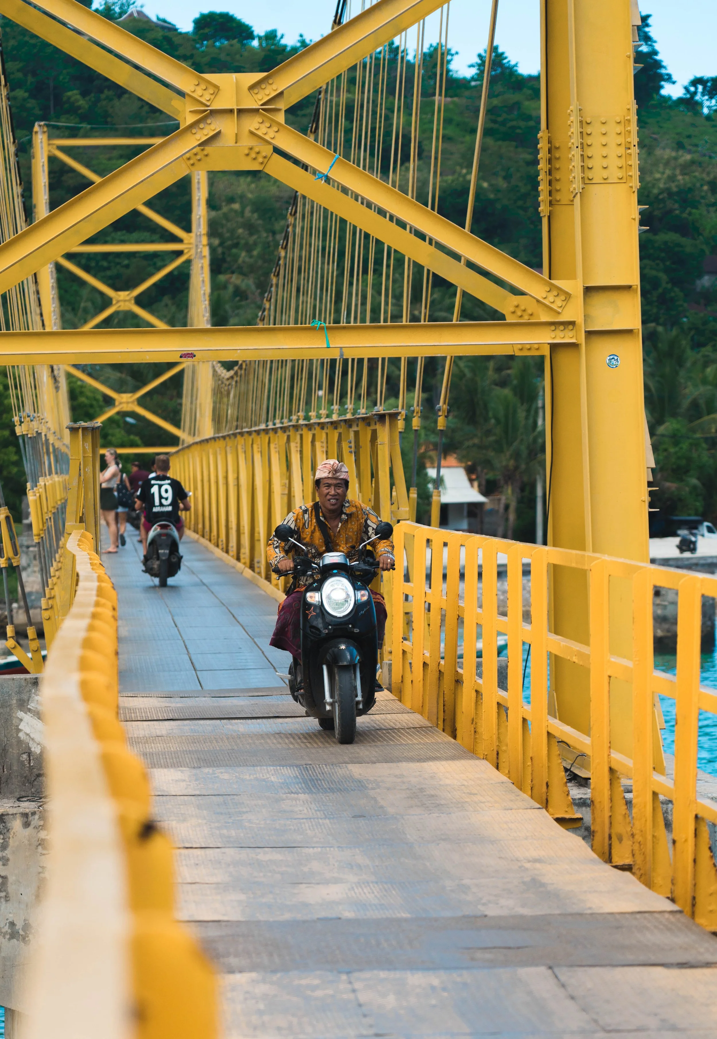 A yellow suspension bridge with motorcycles and pedestrians crossing over it, with a scenic green landscape and trees in the background.