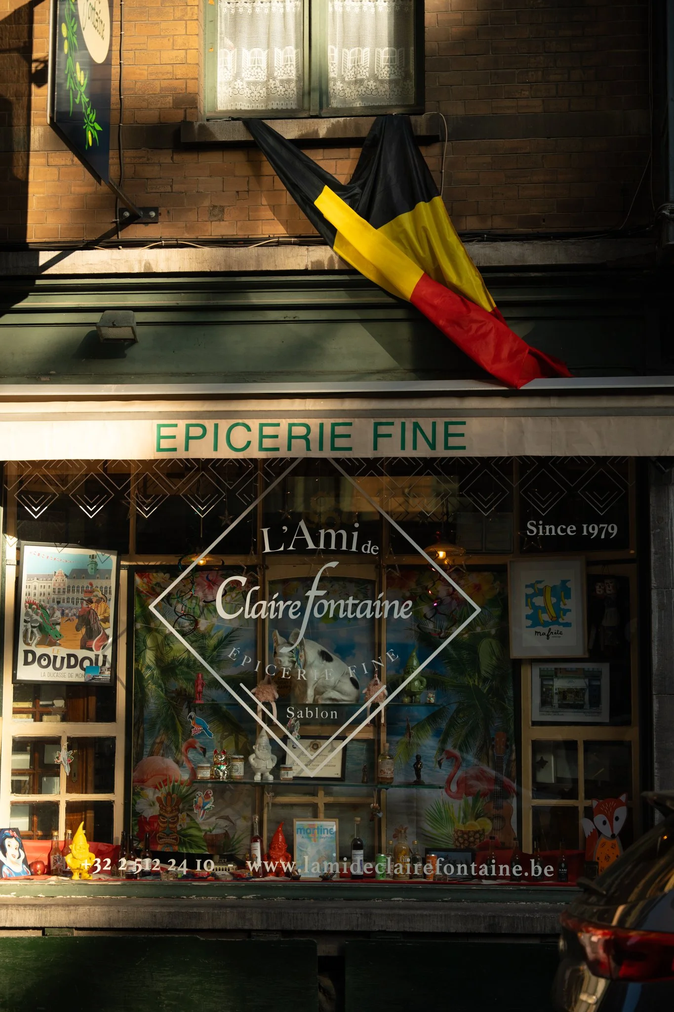 The image shows the storefront of 'Epicerie Fine,' a specialty grocery store. There is a Belgian flag hanging on the wall above the store. The window display features various decorative items, toys, and bottles, along with posters and signs, including the store's name 'Claire Fontaine' and contact information.