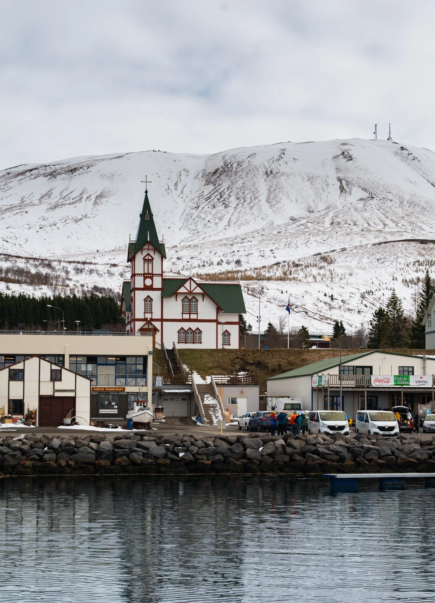 View of a waterfront town with a church, commercial buildings, and parked cars, with a snow-covered hill and communications towers in the background, and a rocky shoreline in the foreground.