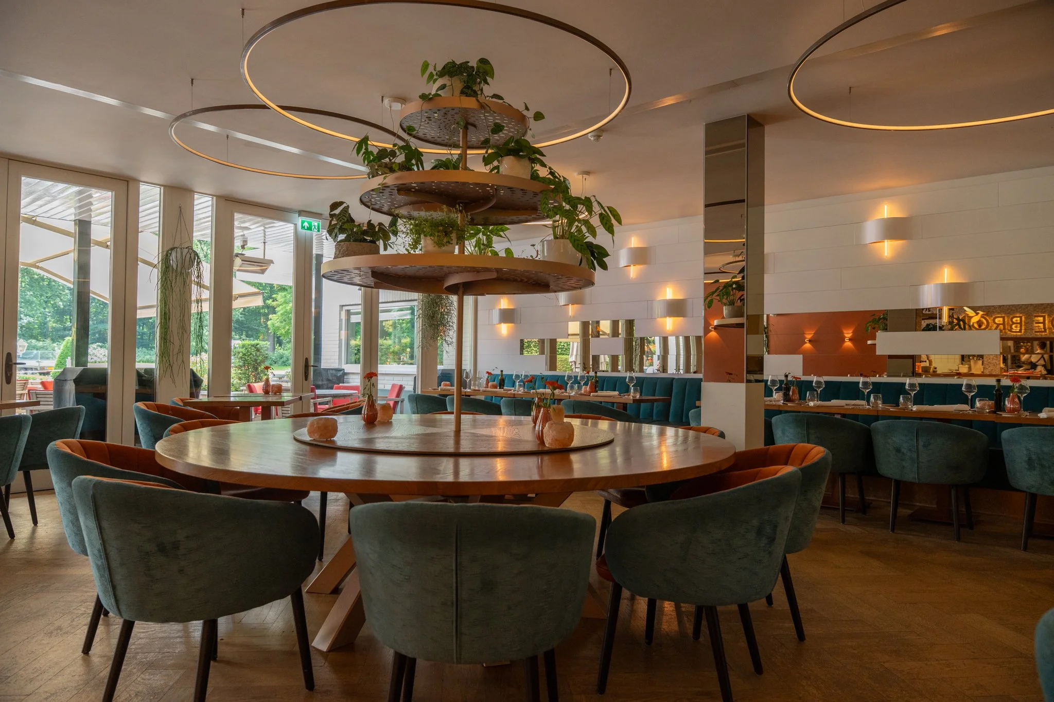 Interieur van een restaurant met grote ronde tafel in het midden, omringd door groene en oranje stoelen, grote ramen aan de buitenkant laten zicht op een groene tuin zien, en binnen goede verlichting en decoraties. Landgoed Groot Warnsborn Arnhem