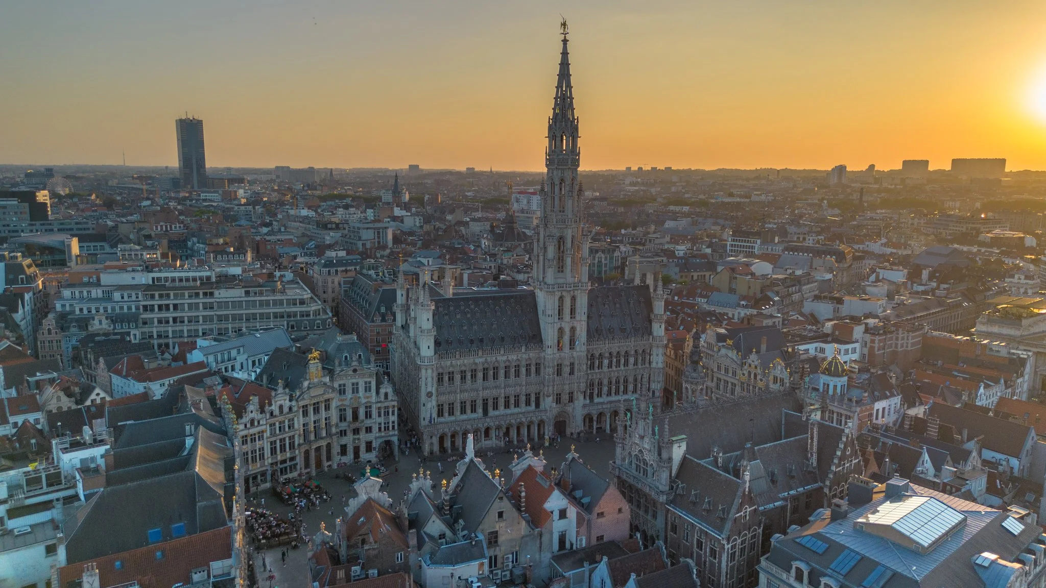 Aerial view of a cityscape during sunset, featuring historic and modern buildings, including a prominent Gothic-style city hall with a tall spire, in a European city.