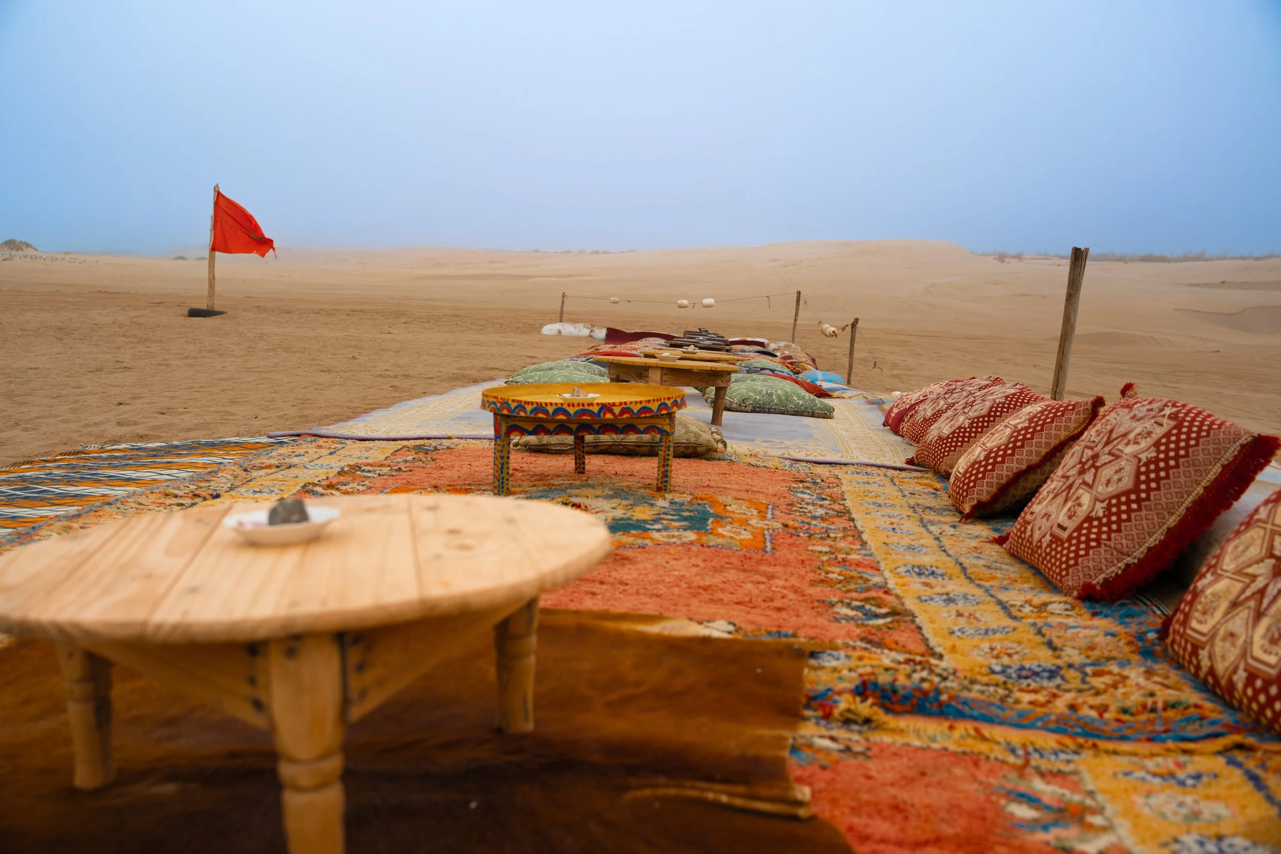A desert scene with colorful patterned rugs and pillows set on the sand, a small round wooden table in the foreground, a red flag on a post to the left, and a makeshift fence with hanging lanterns in the background under a blue sky.