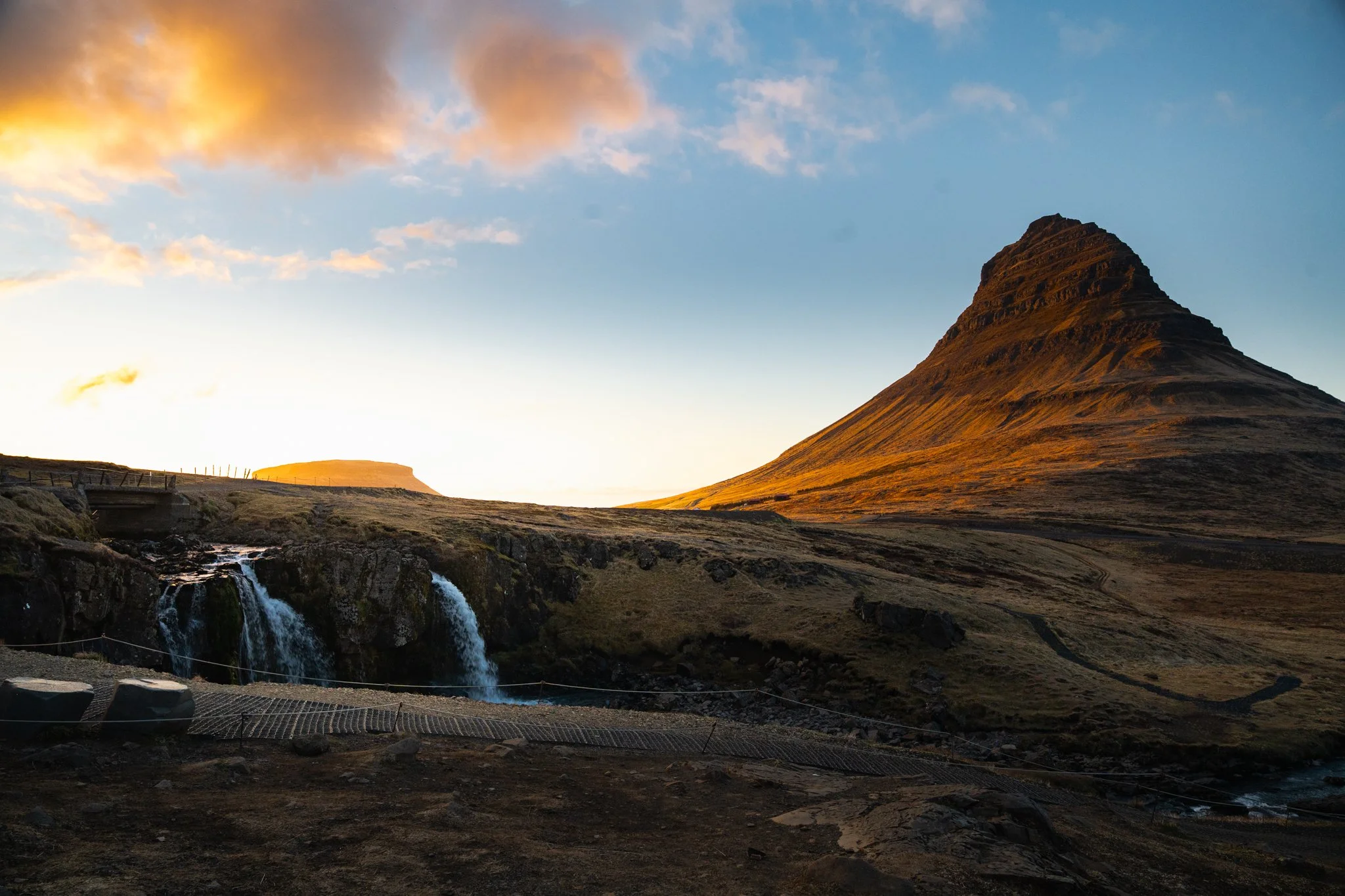 A mountain with a conical shape at sunset, with clouds in the sky and a waterfall in the foreground.