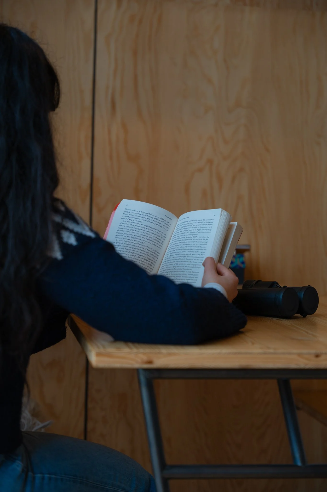 Persoon leest een boek in een houten omgeving, naast een paar verrekijkers op een tafel. Cabiner Groningen, tiny house