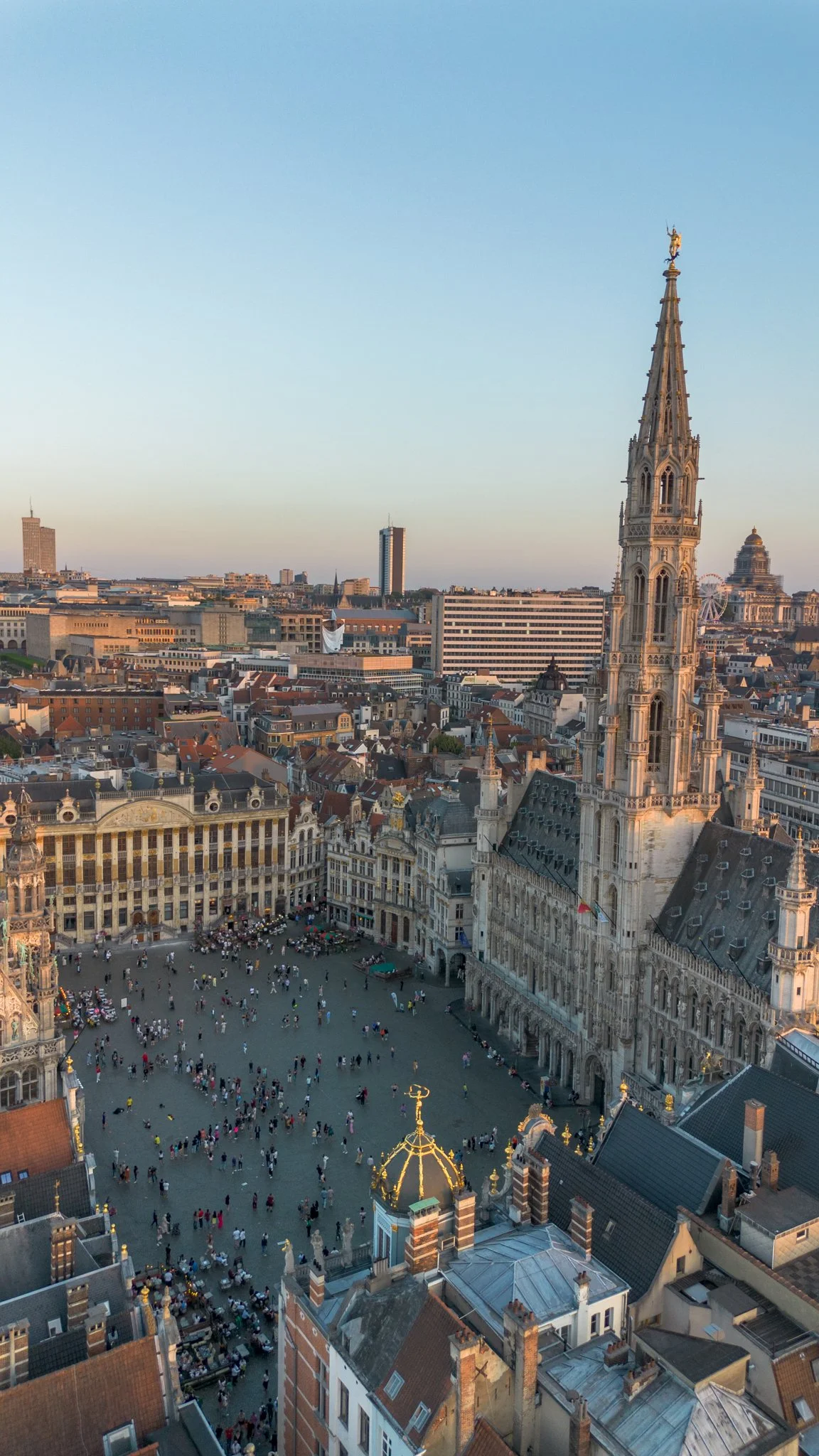 Aerial view of a historic European city square with a large gothic church steeple and bustling crowd, with surrounding buildings and modern cityscape in the background.