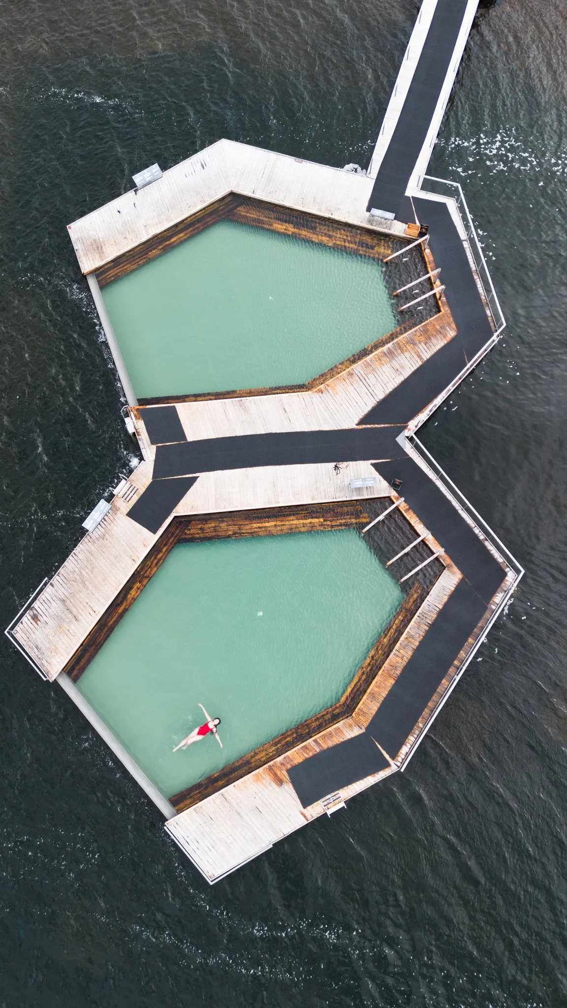 Dronefoto van twee zwembadjes op een houten platform met loopbrug er tussen, gelegen op water, met een persoon die in een van de zwembaden ligt en zich aftekent tegen het water. Vök baths iceland