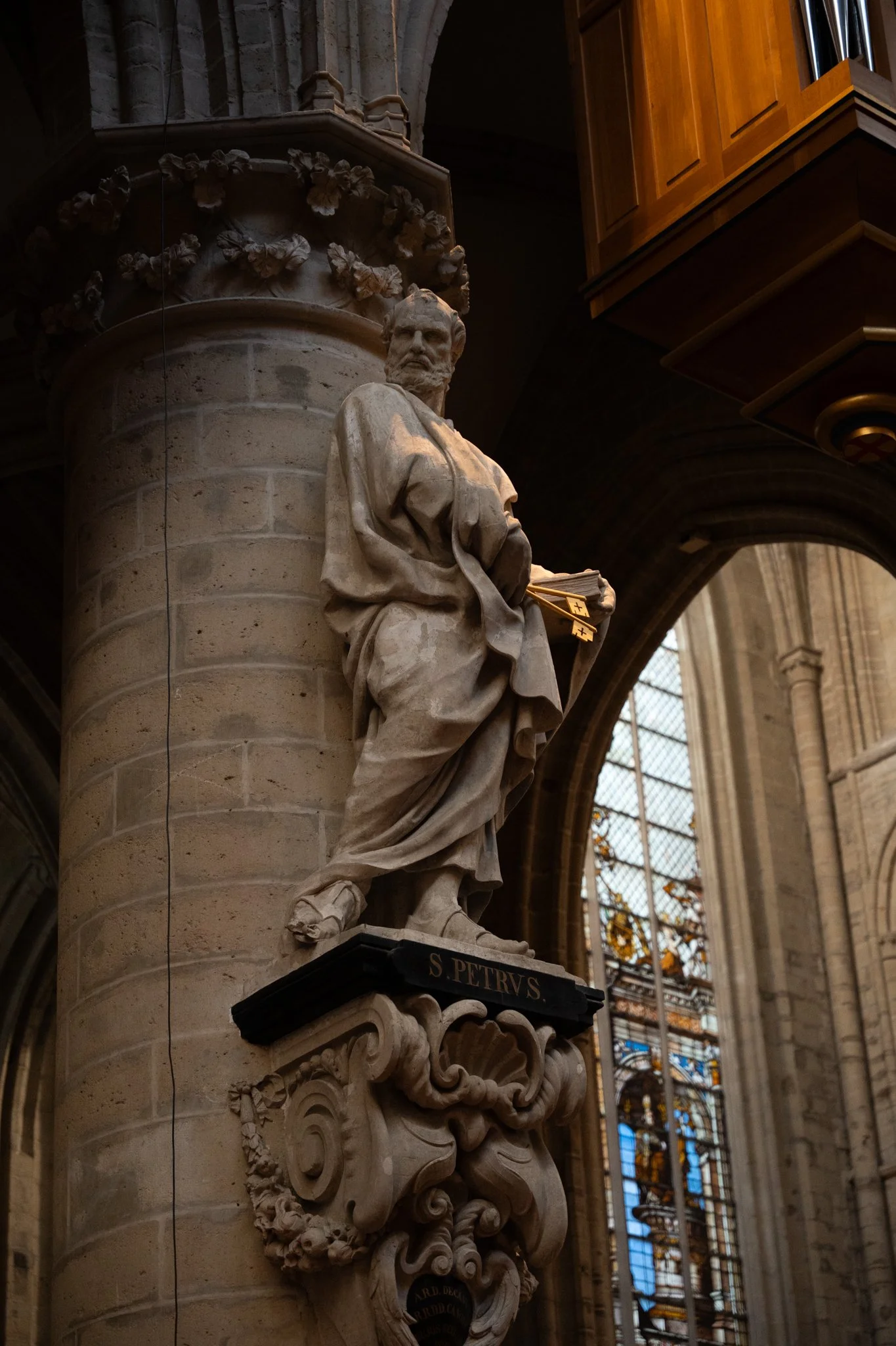 A marble statue of Saint Peter inside a cathedral, holding keys, located on a decorated pedestal labeled "S. PETRVS," with stained glass windows in the background.