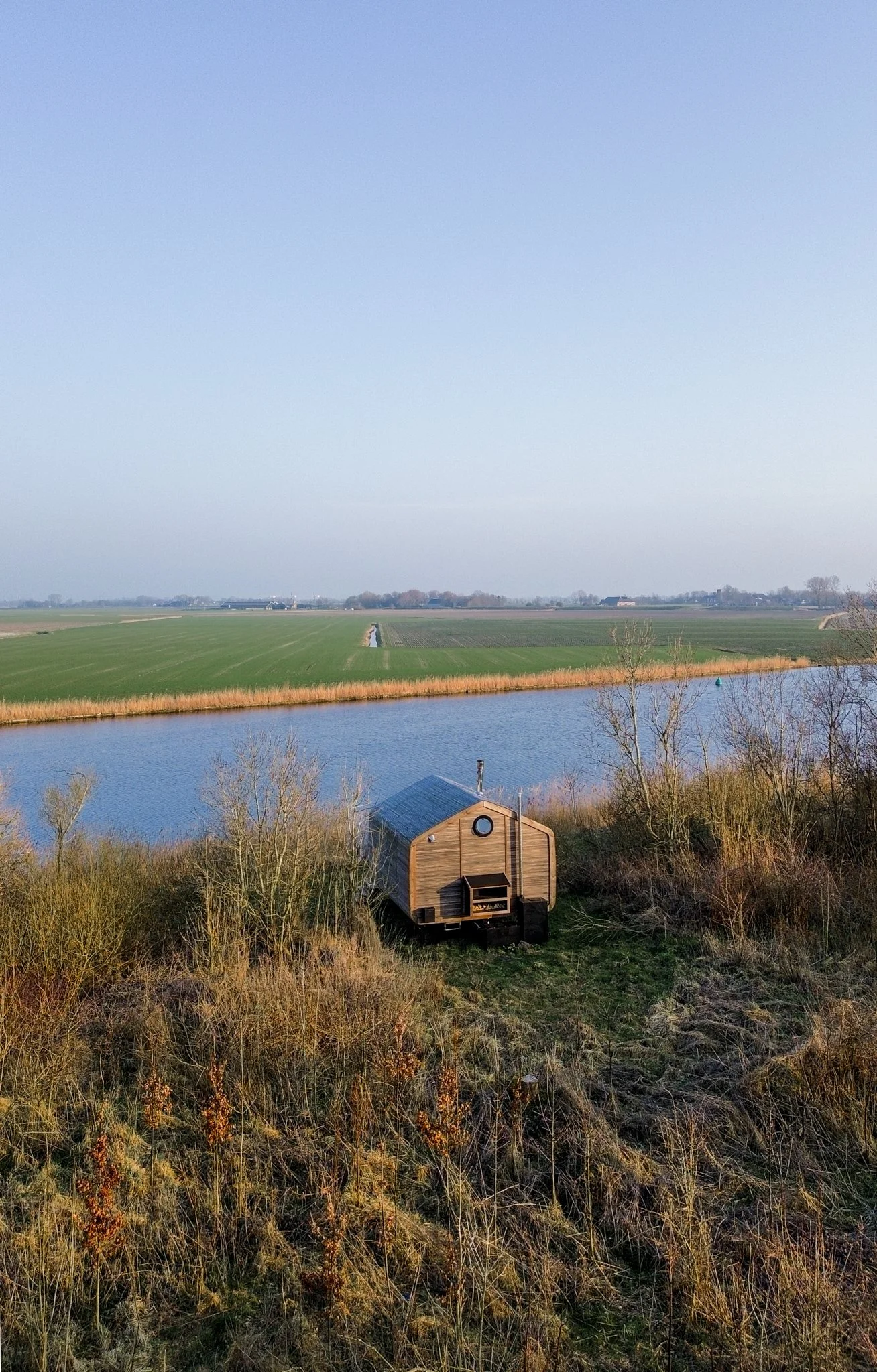 Een houten tiny house naast een rivier in een landelijke omgeving met weilanden en bomen. Cabiner Groningen