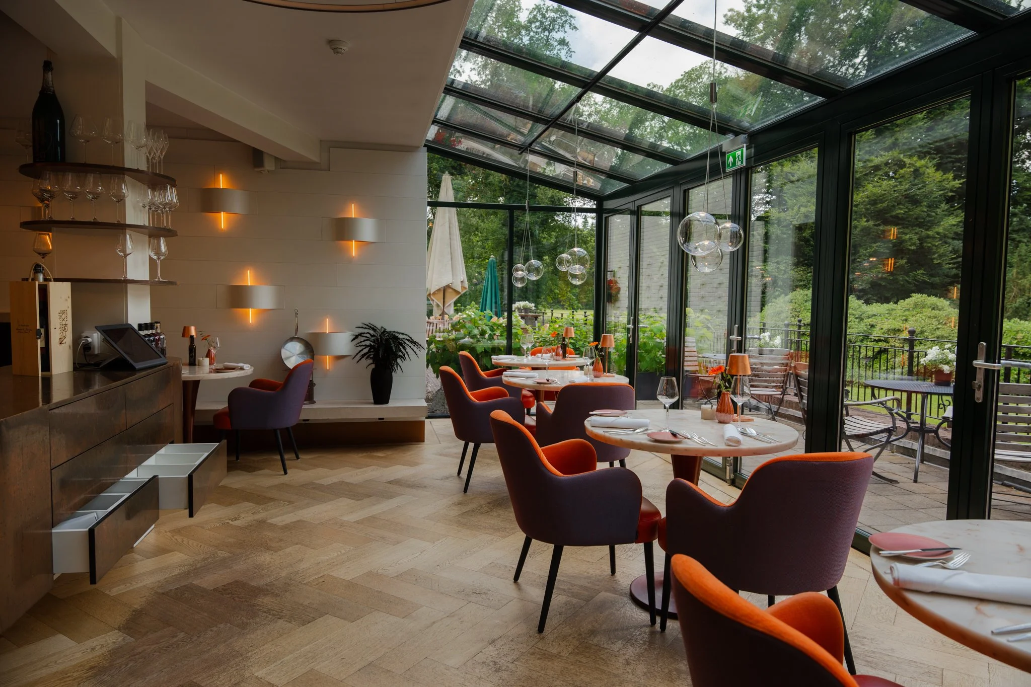 Binnenruimte van een restaurant met grote glazen wanden en een glazen dak, ronde tafels met oranje en paarse stoelen, decoratieve hanglampen en uitzicht op een groene tuin buiten. Landgoed Groot Warnsborn in Arnhem