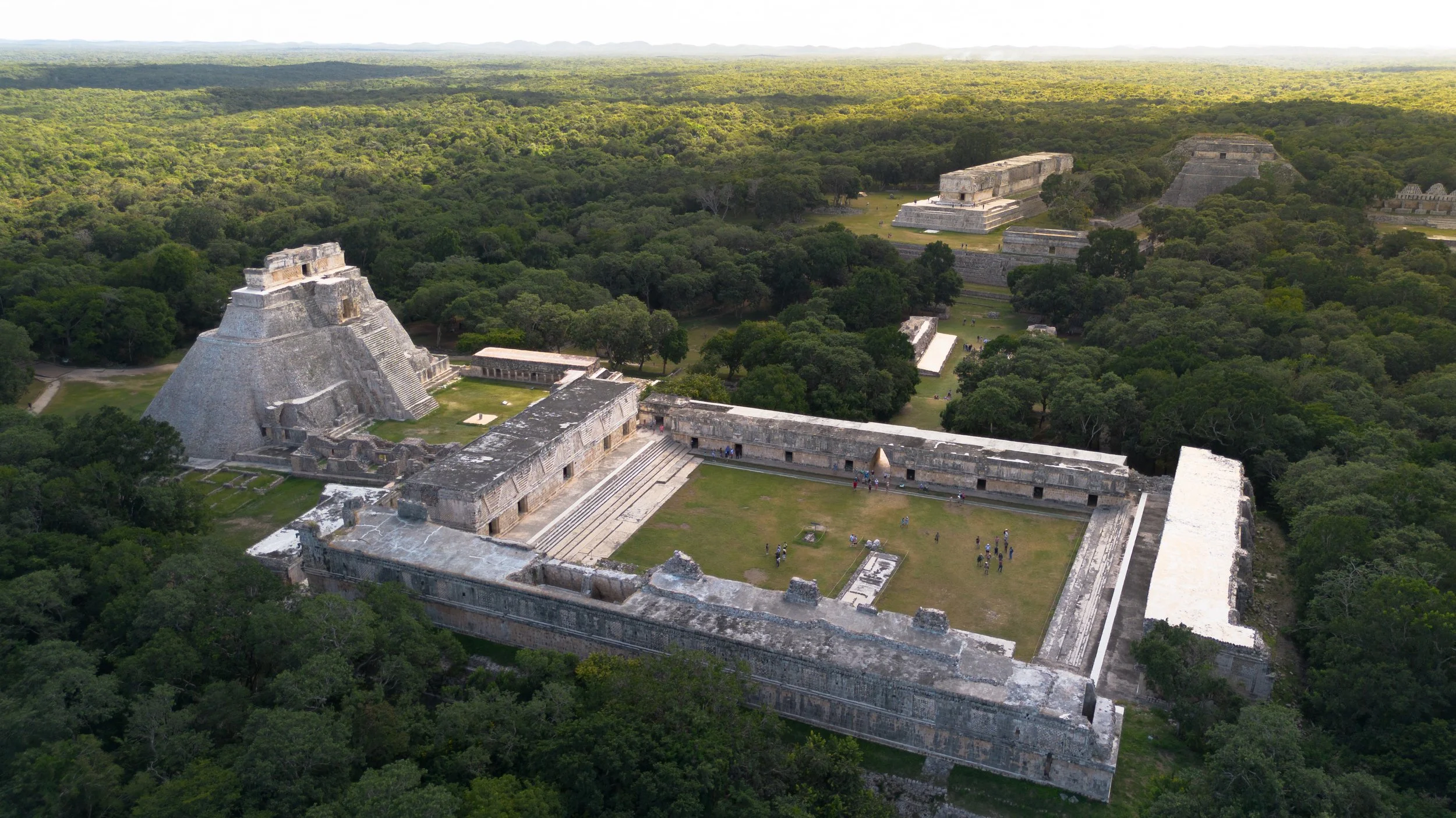An aerial view of ancient Mayan ruins surrounded by dense green jungle with multiple large stone pyramids and temples.