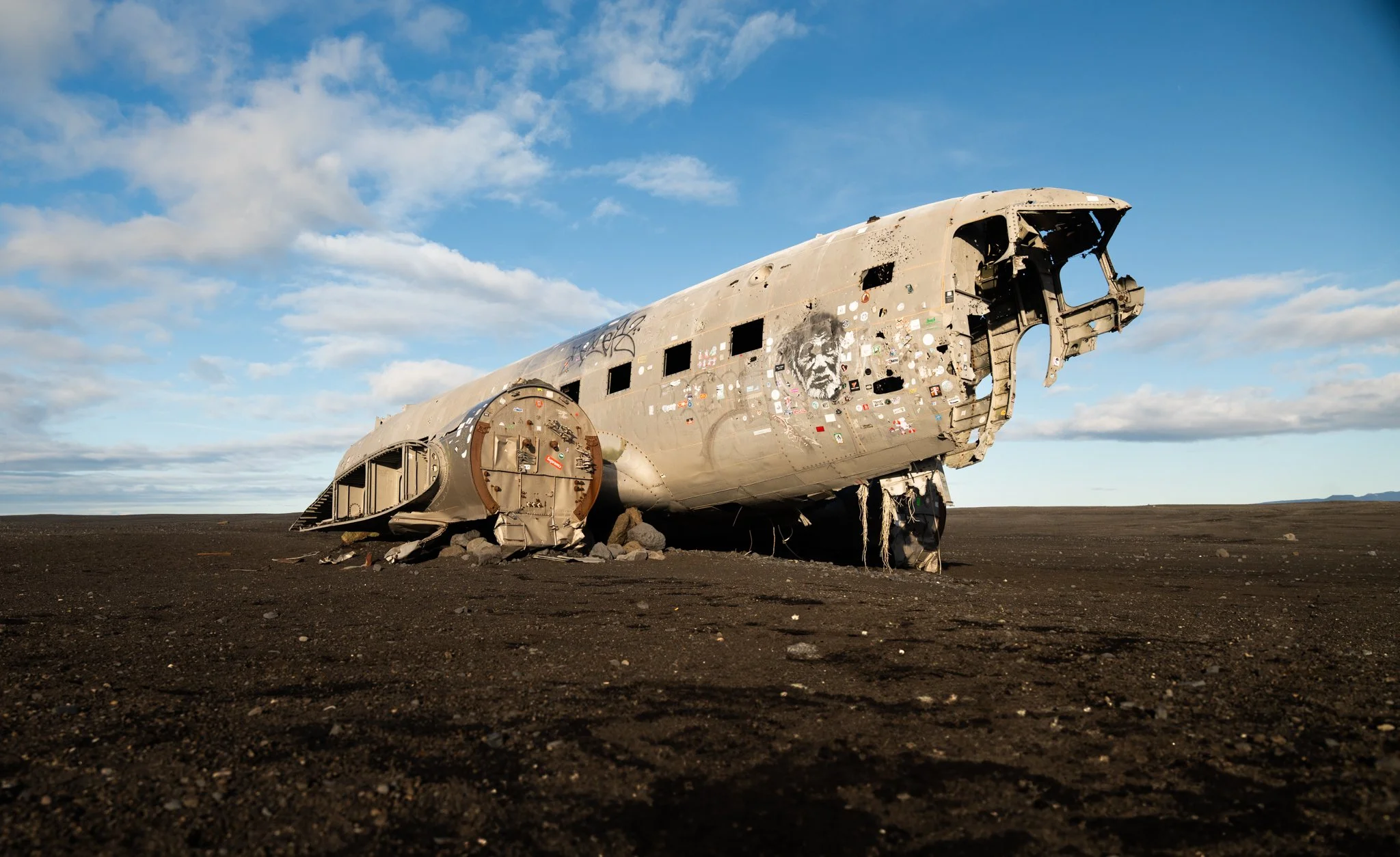 A crashed airplane fuselage lying on barren land, covered with graffiti and stickers, under a partly cloudy sky.