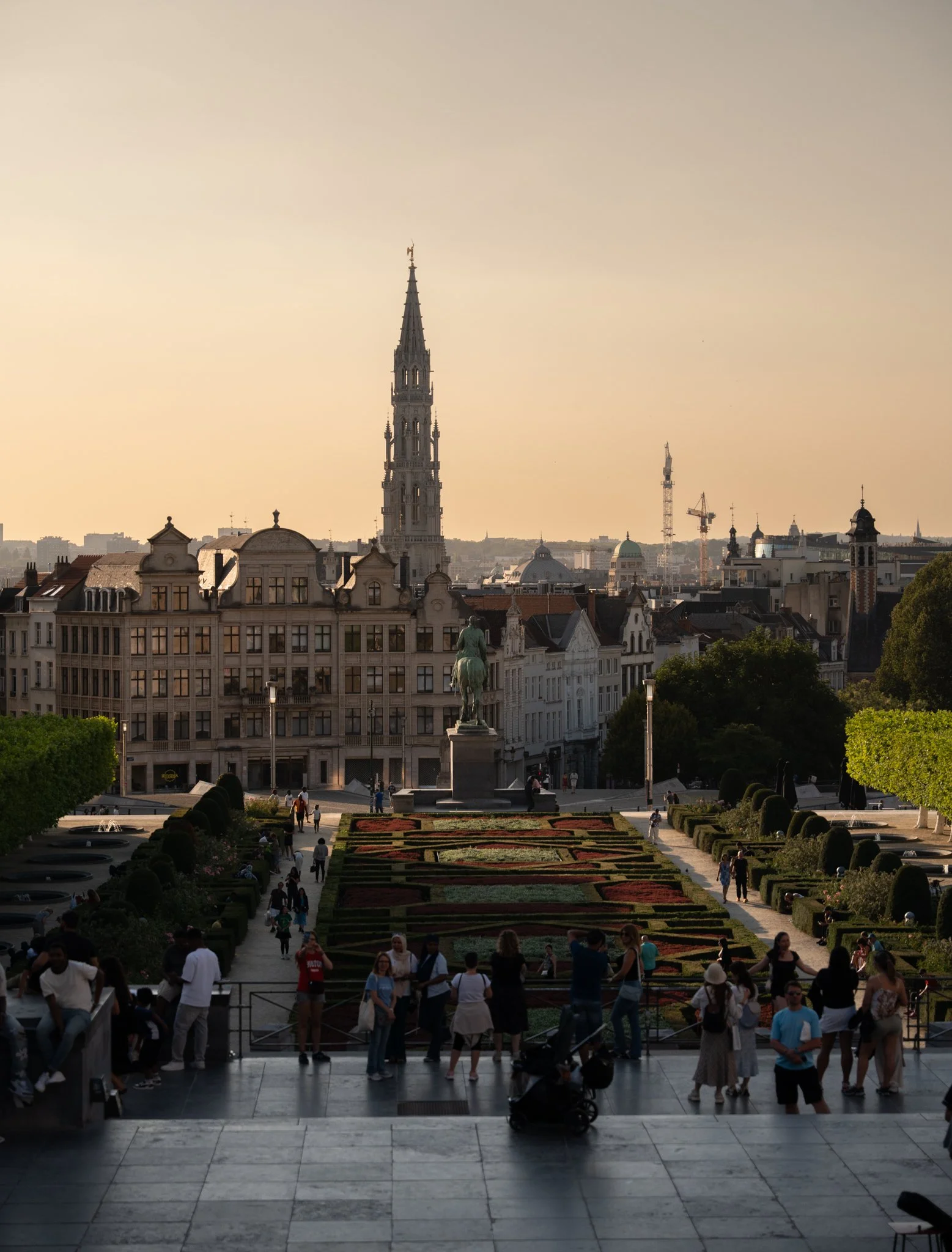 View of a city plaza with a staircase leading up to a park, with people walking and gathering, and a statue of a person on horseback in the foreground. In the background, historic buildings and a church steeple are visible, with construction cranes and a tall church spire against the sunset sky.