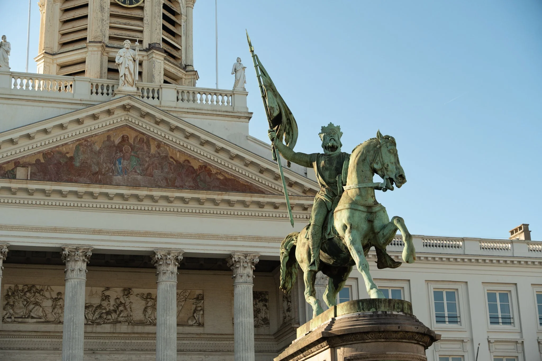 Statue of a man on a horse, holding a flag, in front of a classical building with statues and detailed architectural elements.