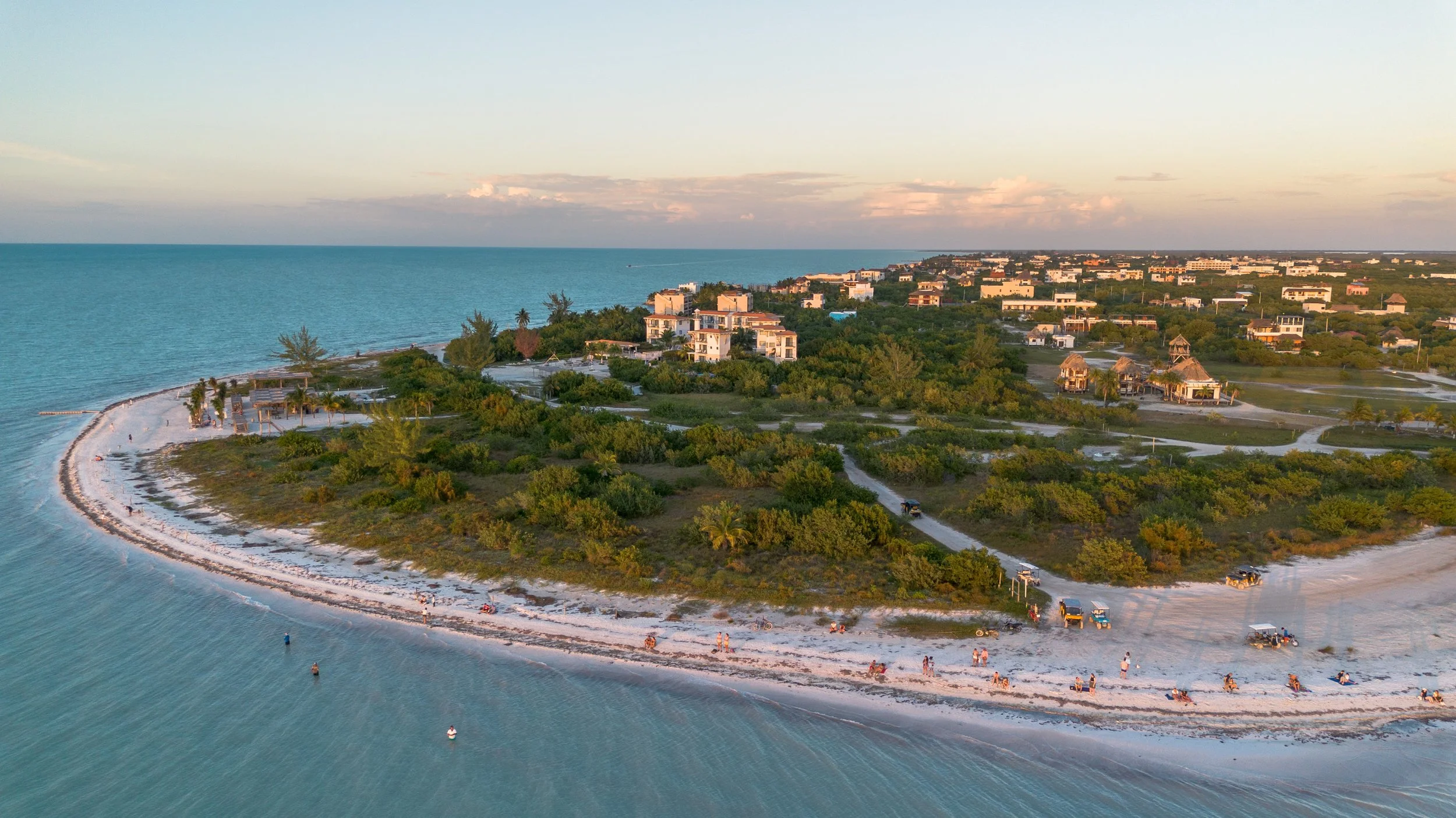 Aerial view of a beachside resort with white sandy beaches, green vegetation, and buildings, near the ocean with a curved shoreline during sunset.