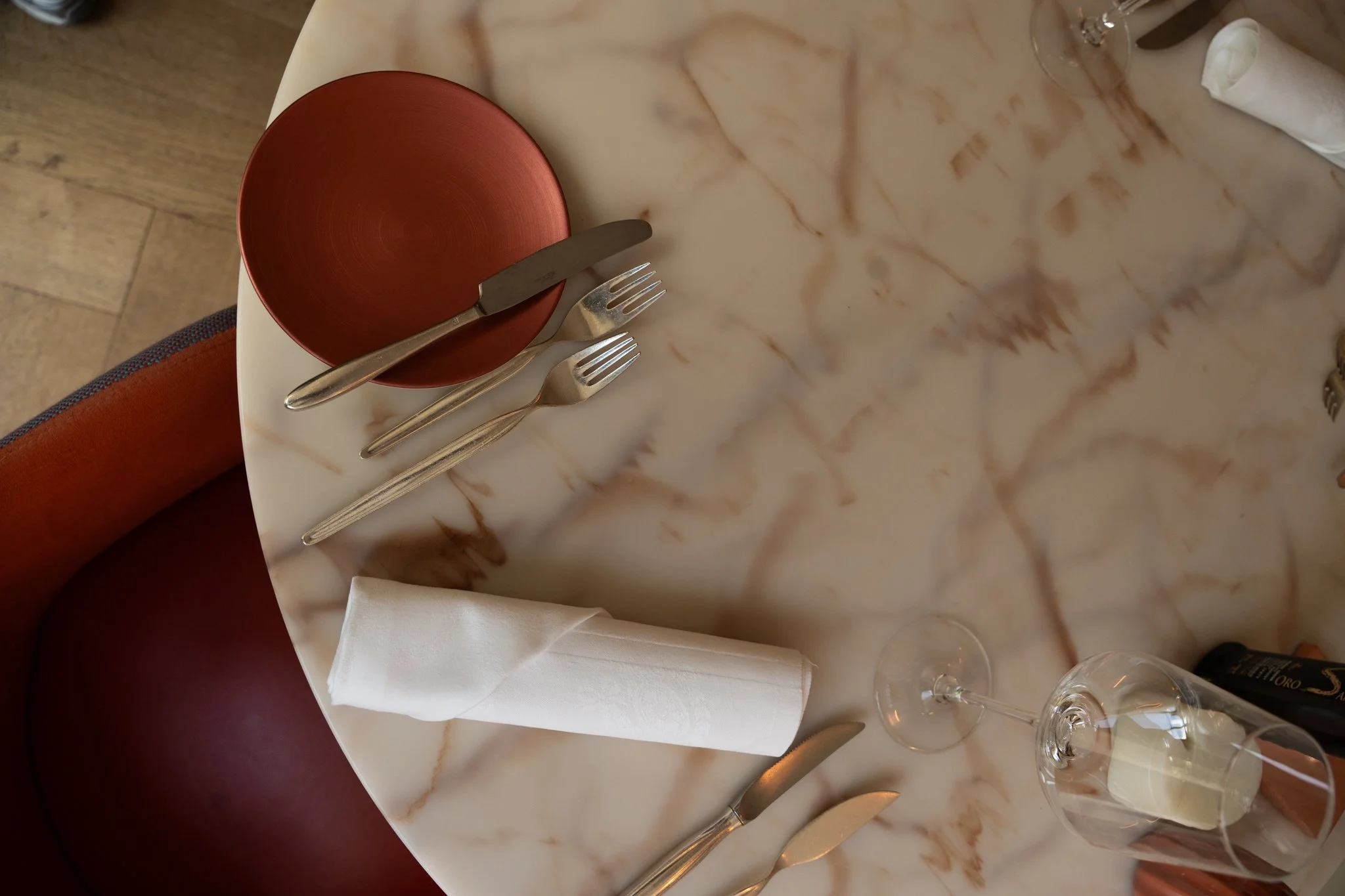 Een ronde tafel met een witte marmeren top, een rood bord, een mes, twee vorken, een wit opgerolde doek, twee wijn glazen en een flesje Brasserie De Bron Landgoed groot warnsborn Arnhem