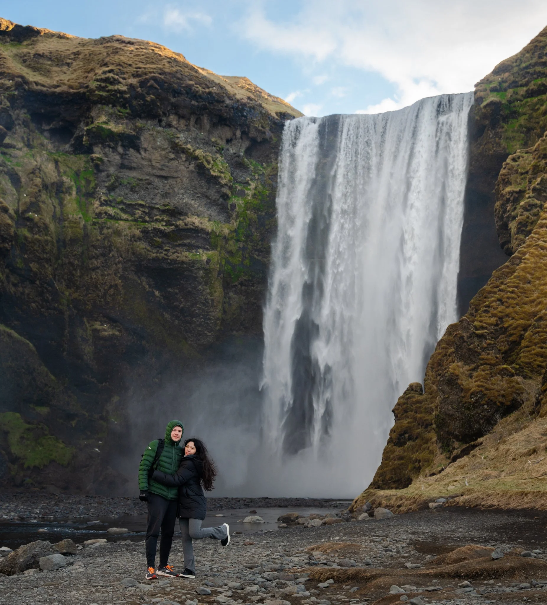 Twee mensen staan bij een waterval in Ijsland. omringd door rotsen en groene heuvels, onder een gedeeltelijk bewolkte blauwe hemel. Skogafoss