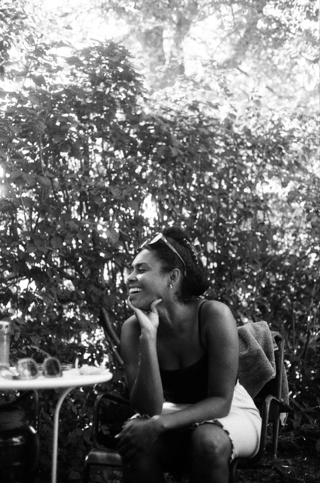 A woman sitting outdoors, smiling and looking to the side, with a table holding a water bottle and sunglasses in front of her, surrounded by leafy trees.
