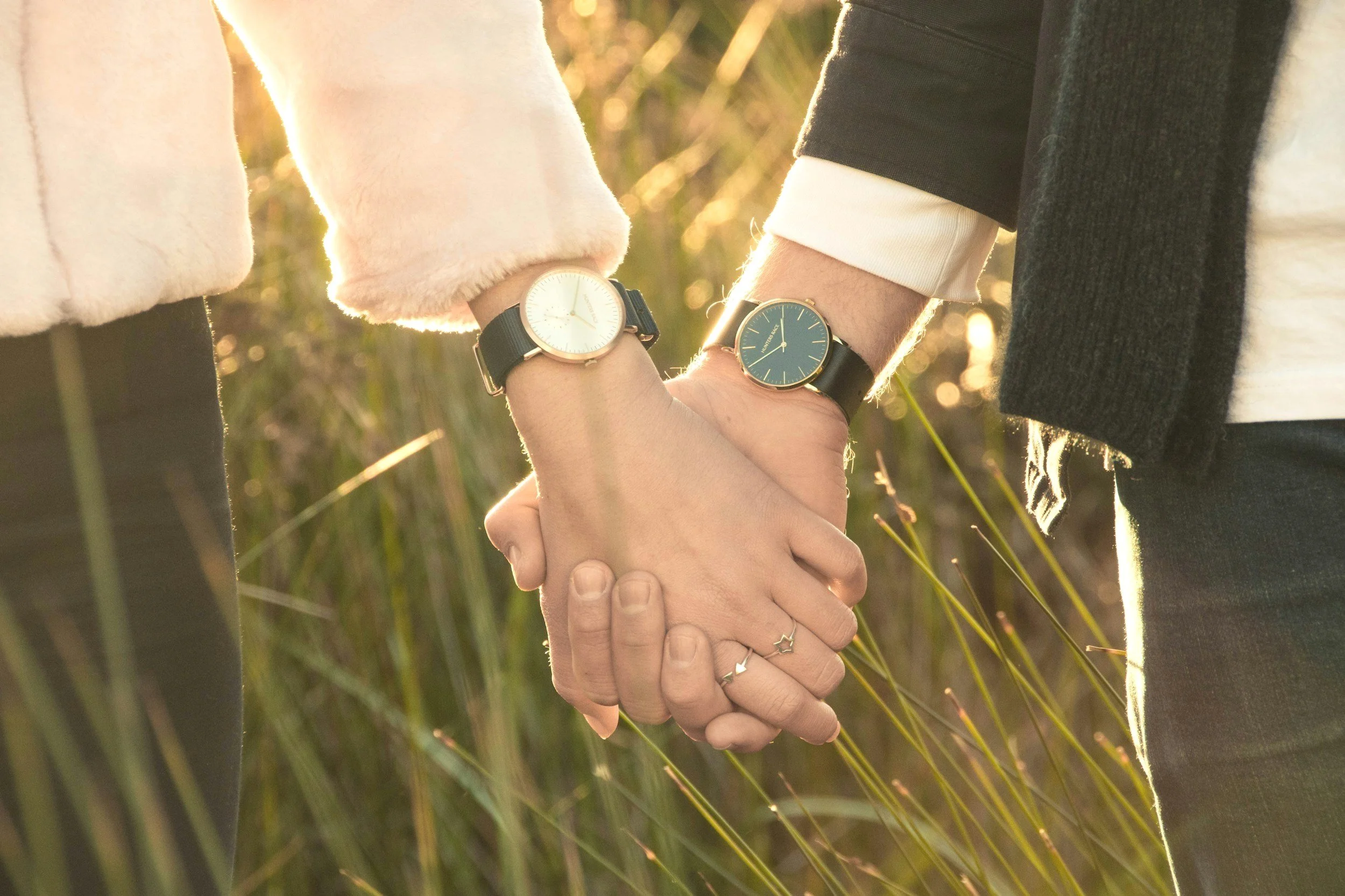 Close-up of two people holding hands outdoors in a grassy area, wearing watches, with sunlight in the background.