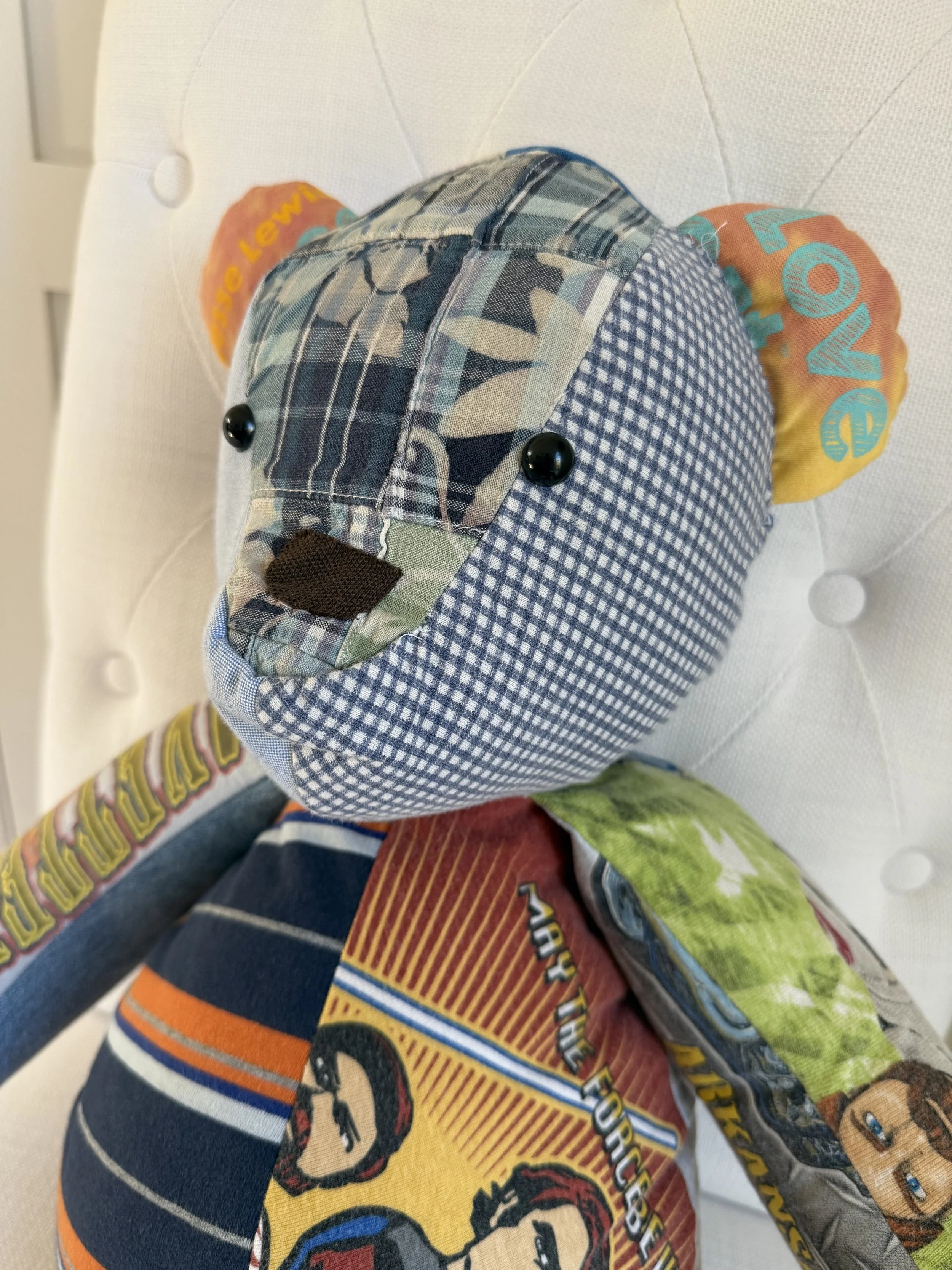 A fabric patchwork dog plush toy with button eyes, made from camo and checkered fabric, sitting on a white tufted cushioned surface.