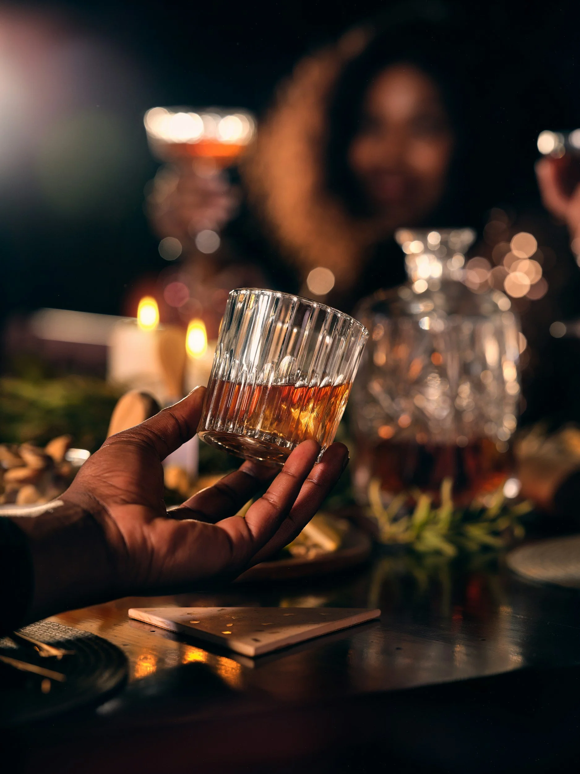 Person holding a glass of whiskey at a dinner or celebration with a woman in the background, candles and glass decanter on the table.