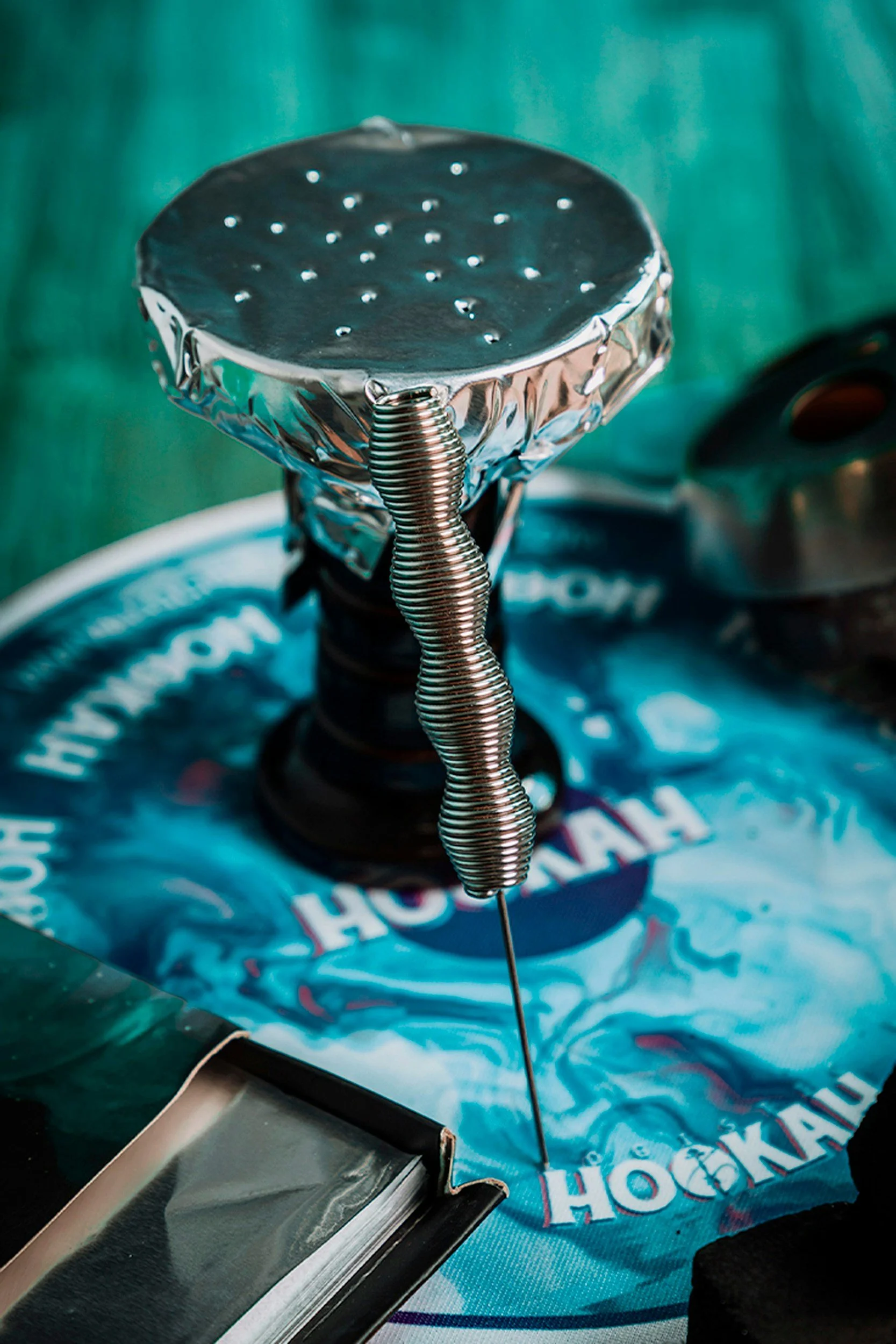 Close-up of a hookah on a table with a teal background, featuring a metallic bowl with small holes, a coil-wrapped hose, and a black base, with a blue Hokah branded napkin underneath.