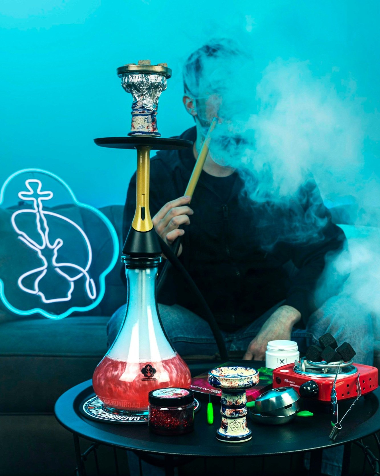 Person sitting on sofa smoking hookah with a table of hookah supplies and accessories, illuminated by neon signs and blue lighting.