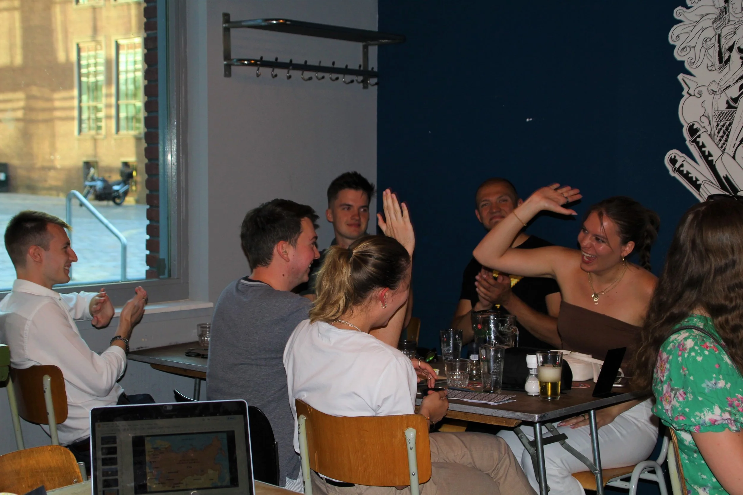 Group of friends enjoying an EESA celebration at a restaurant, raising their hands for a high five, with smiles on their faces, glasses on the table, and a window showing a street scene outside.