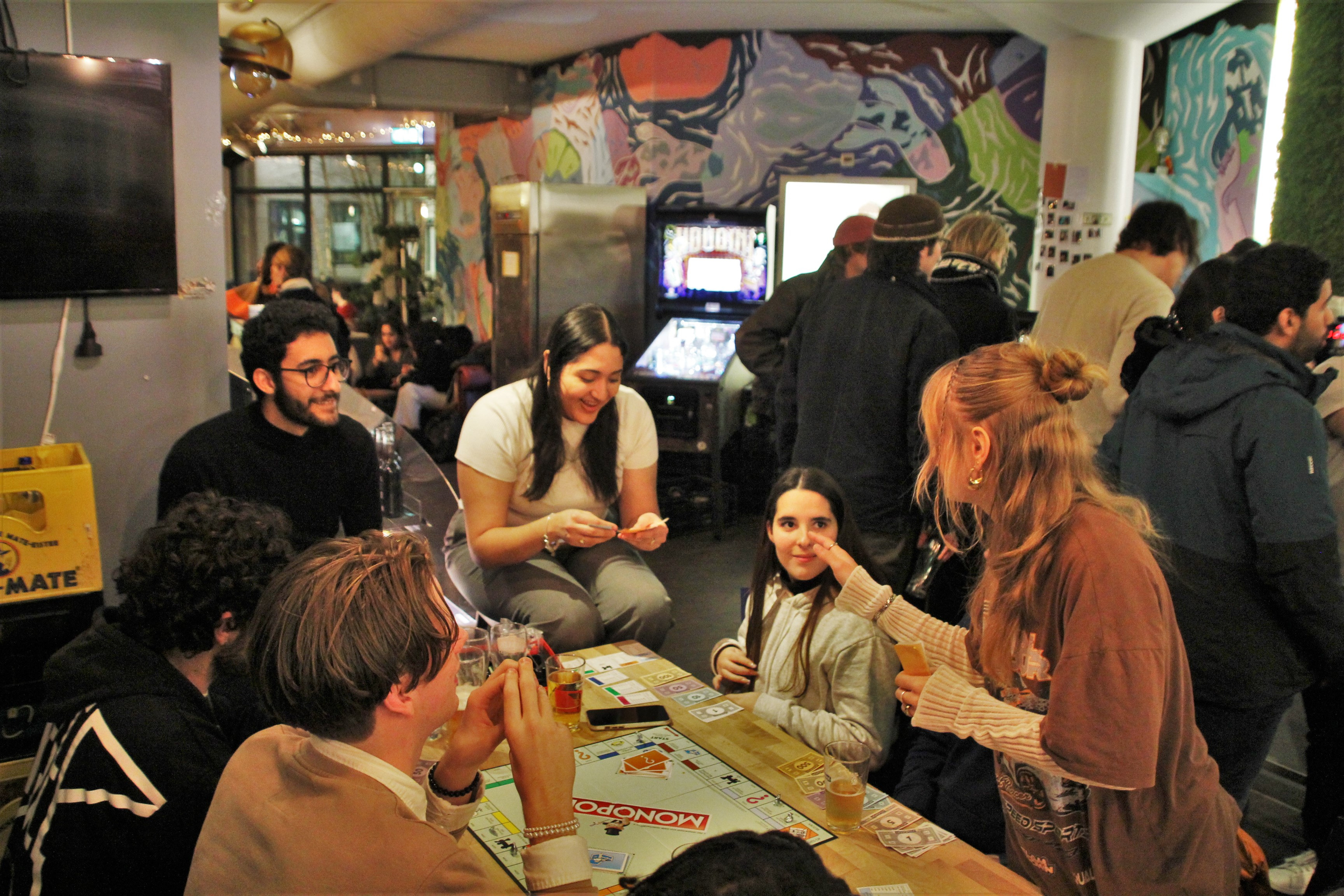 A group of people playing Monopoly at a table in a lively, colorful cafe or restaurant, with some standing and some sitting, and a mural on the wall behind them.