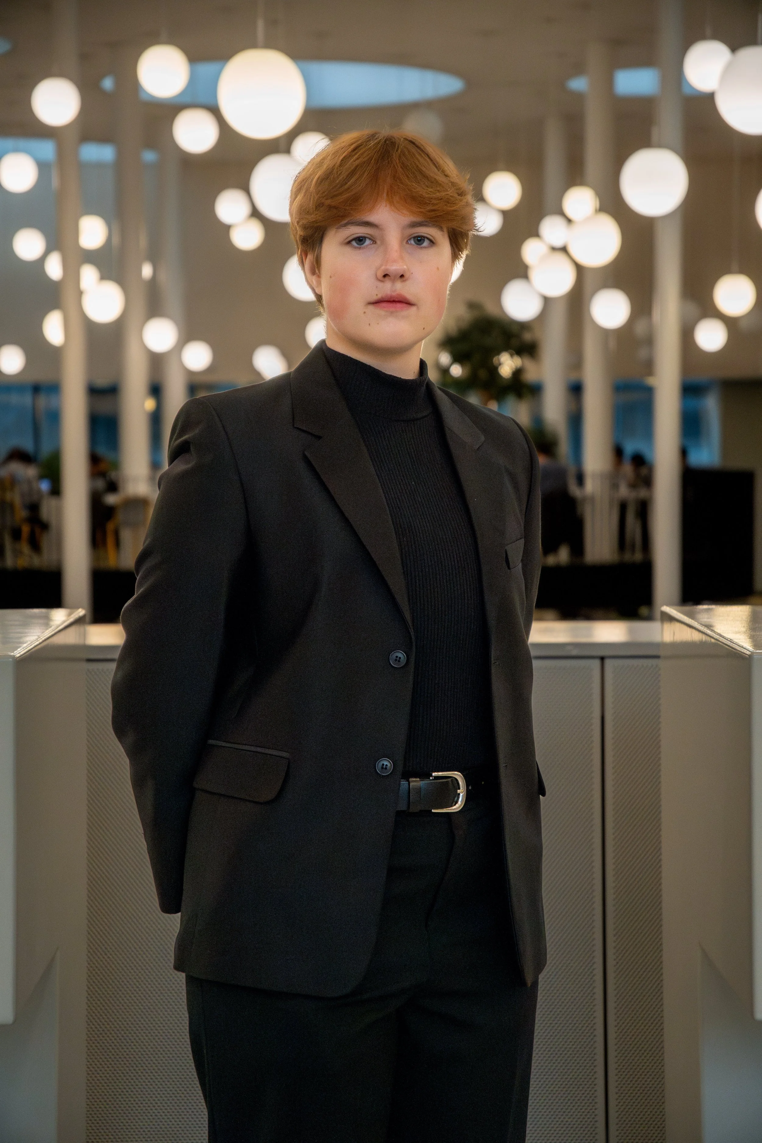 A young male student with short red hair, dressed in a black suit, standing in a modern indoor space with hanging round lights in the background.