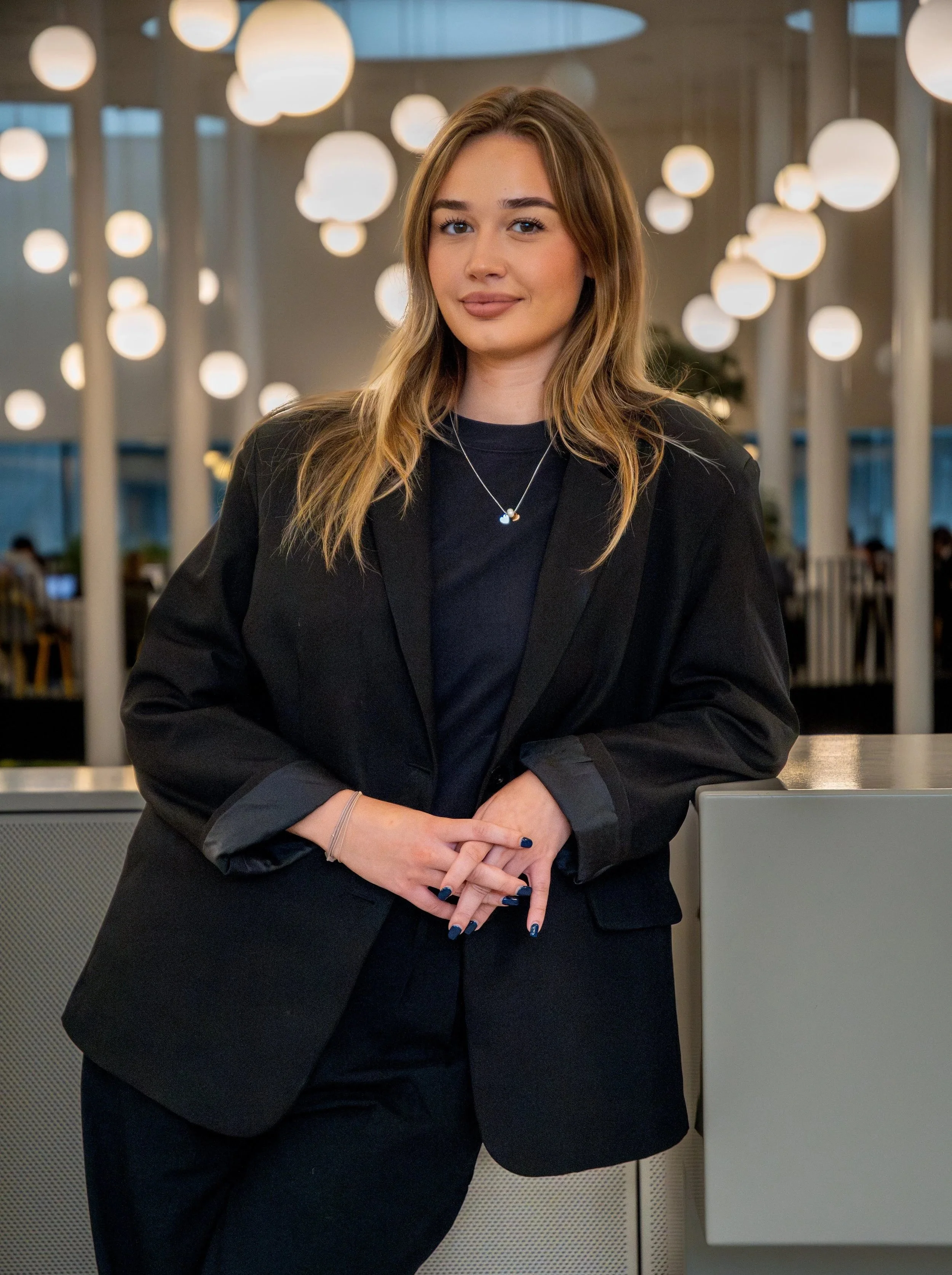 A young female student with long, wavy, light brown hair standing in a modern, well-lit indoor space, wearing a black blazer over a black top, with her hands clasped in front of her.