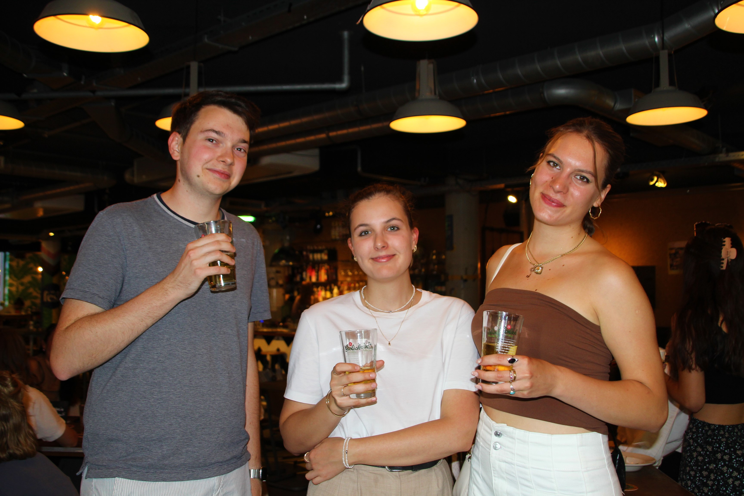 Three young Eastern European adults standing in a bar or restaurant, holding glasses of drinks and smiling at the camera, with warm lighting and a bar in the background.