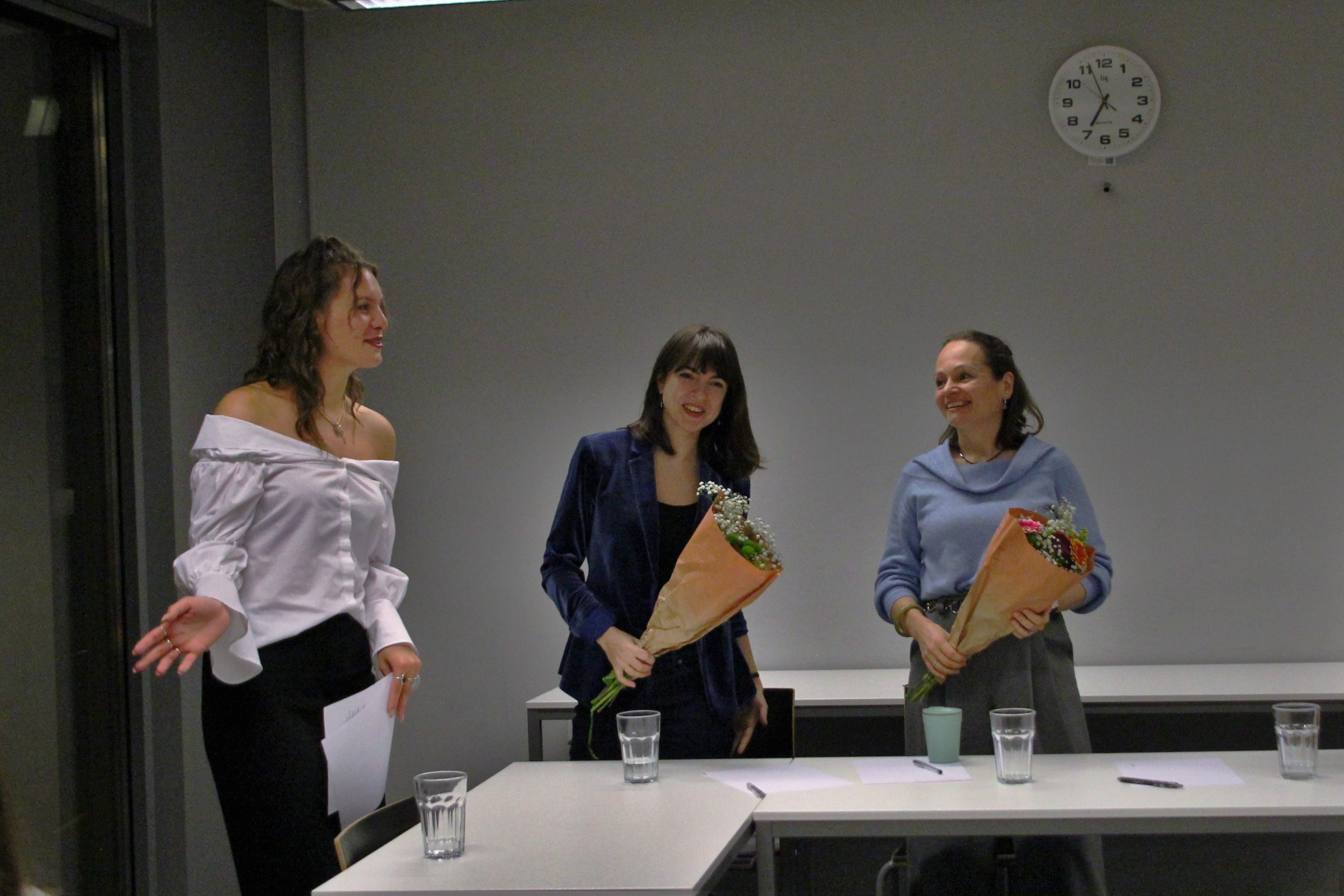 Three women standing behind a conference table, each holding a bouquet of flowers, smiling and engaging in conversation during a formal event in a room with a clock on the wall.